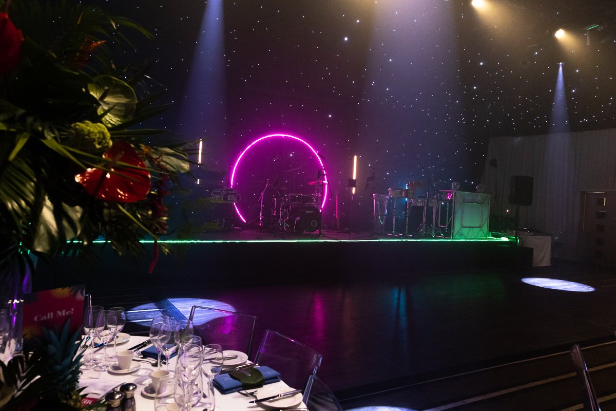 Empty stage decorated with neon pink and green lights, a drum set, and electronic equipment, with a starry ceiling and a floral arrangement in the foreground at a party or event venue.