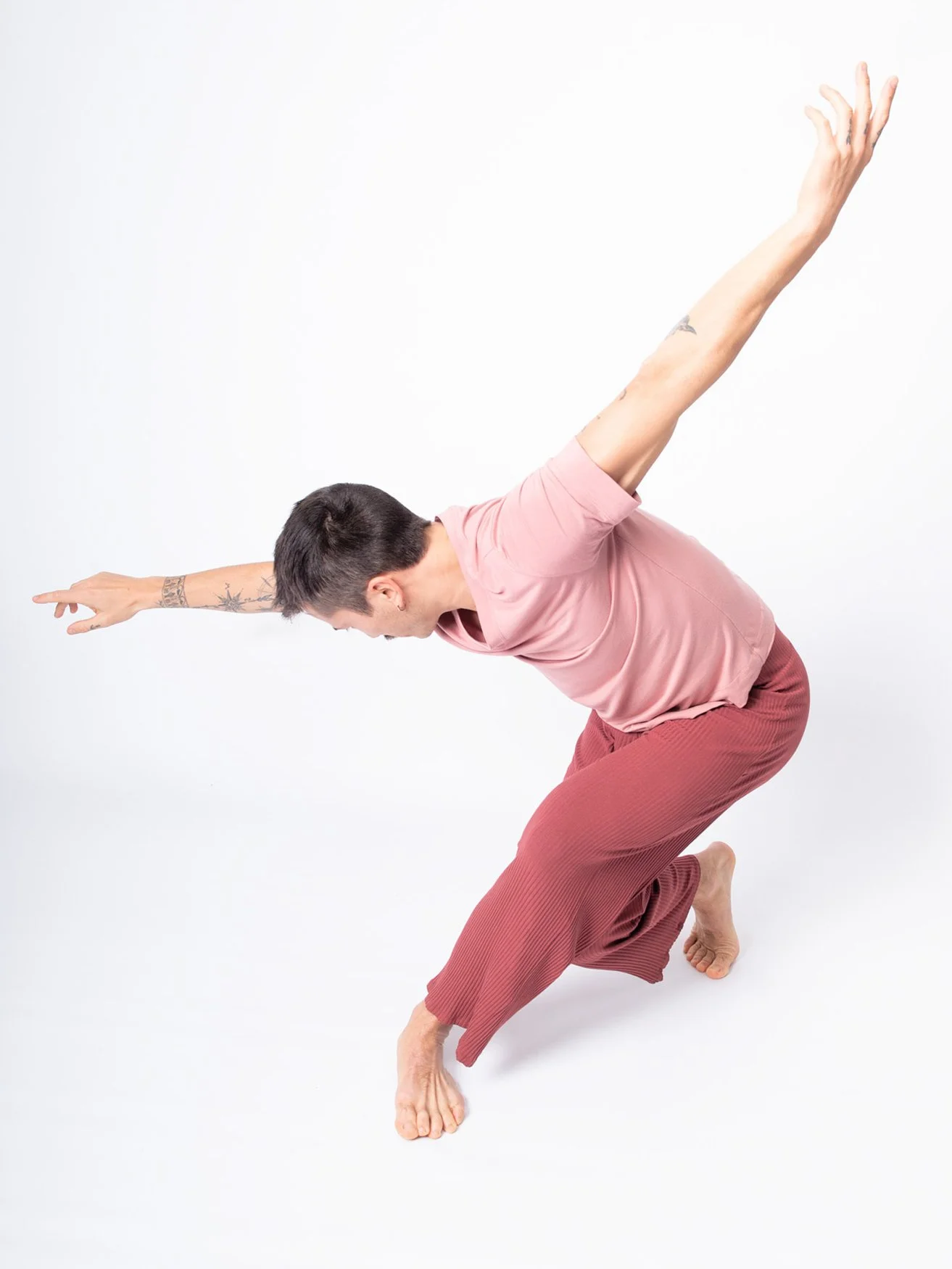 Une femme faisant une pose de yoga ou de méditation, assise en équilibre sur un pied, avec les bras étendus en arrière, vêtue d'un t-shirt rose et pantalon marron. Elle est inclinée en avant, avec la tête baissée.