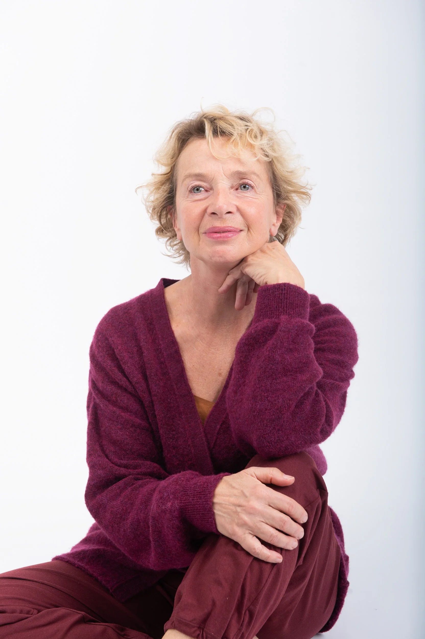Une femme âgée assise, portant un pull violet et un pantalon marron, avec des cheveux bouclés blonds, regardant pensive contre un fond blanc.