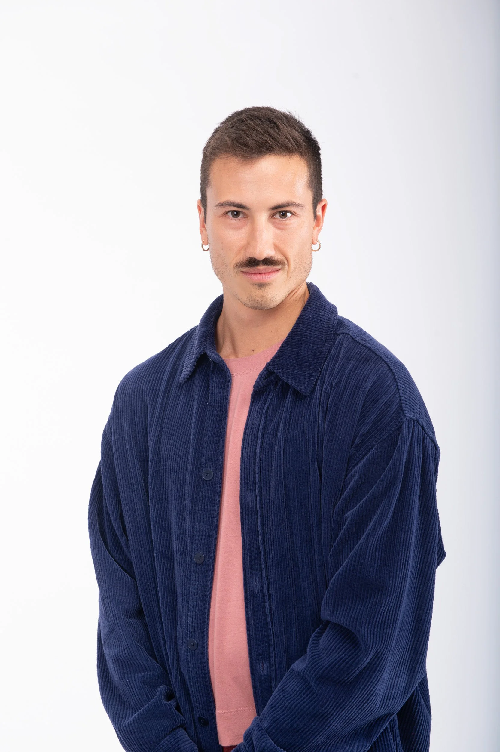Jeune homme avec cheveux courts, moustache, boucles d'oreilles, portant une veste en velours bleu et un t-shirt rose, posé contre un fond blanc.