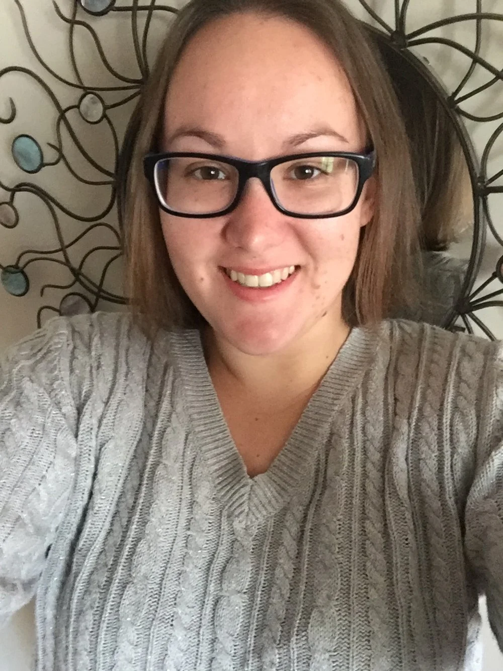 A woman with glasses taking a selfie, smiling, lying on a bed with a decorative metal headboard in the background.