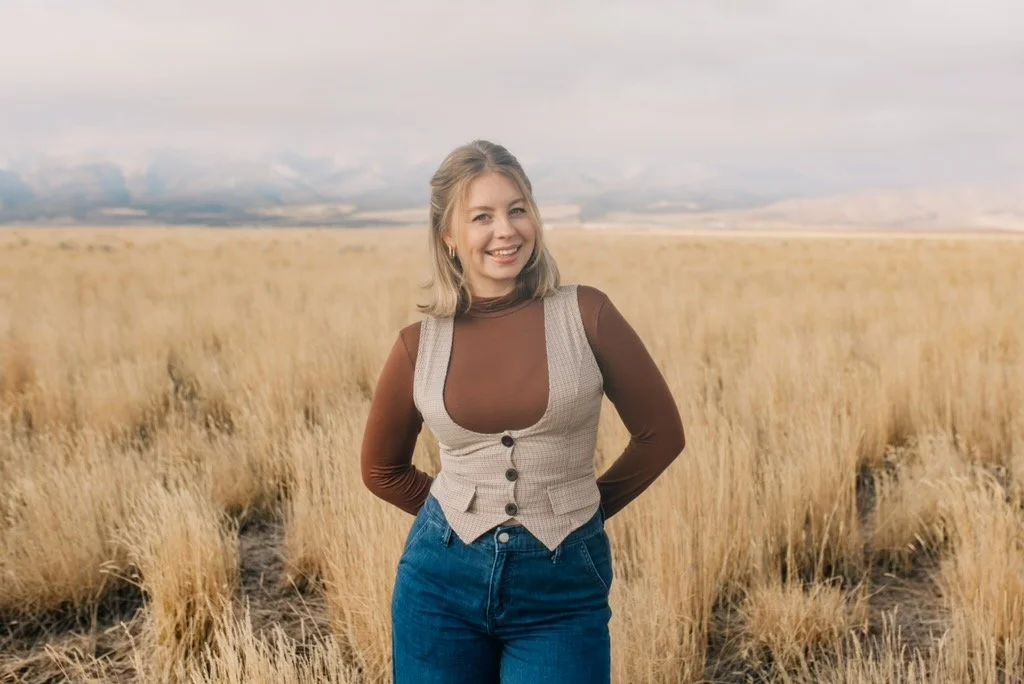 A woman with shoulder-length blonde hair smiling in a field of tall yellow grass with mountains in the background.