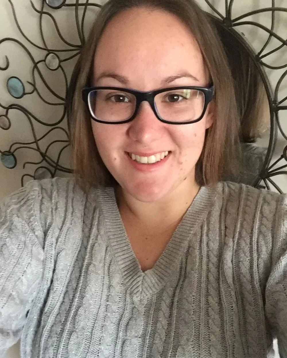 A woman with shoulder-length brown hair, wearing glasses and a light gray cable-knit sweater, smiling at the camera with a decorative metal wall art behind her.