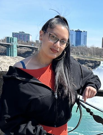 Woman with glasses and long dark hair standing outdoors near water, with city buildings and a bridge in the background.