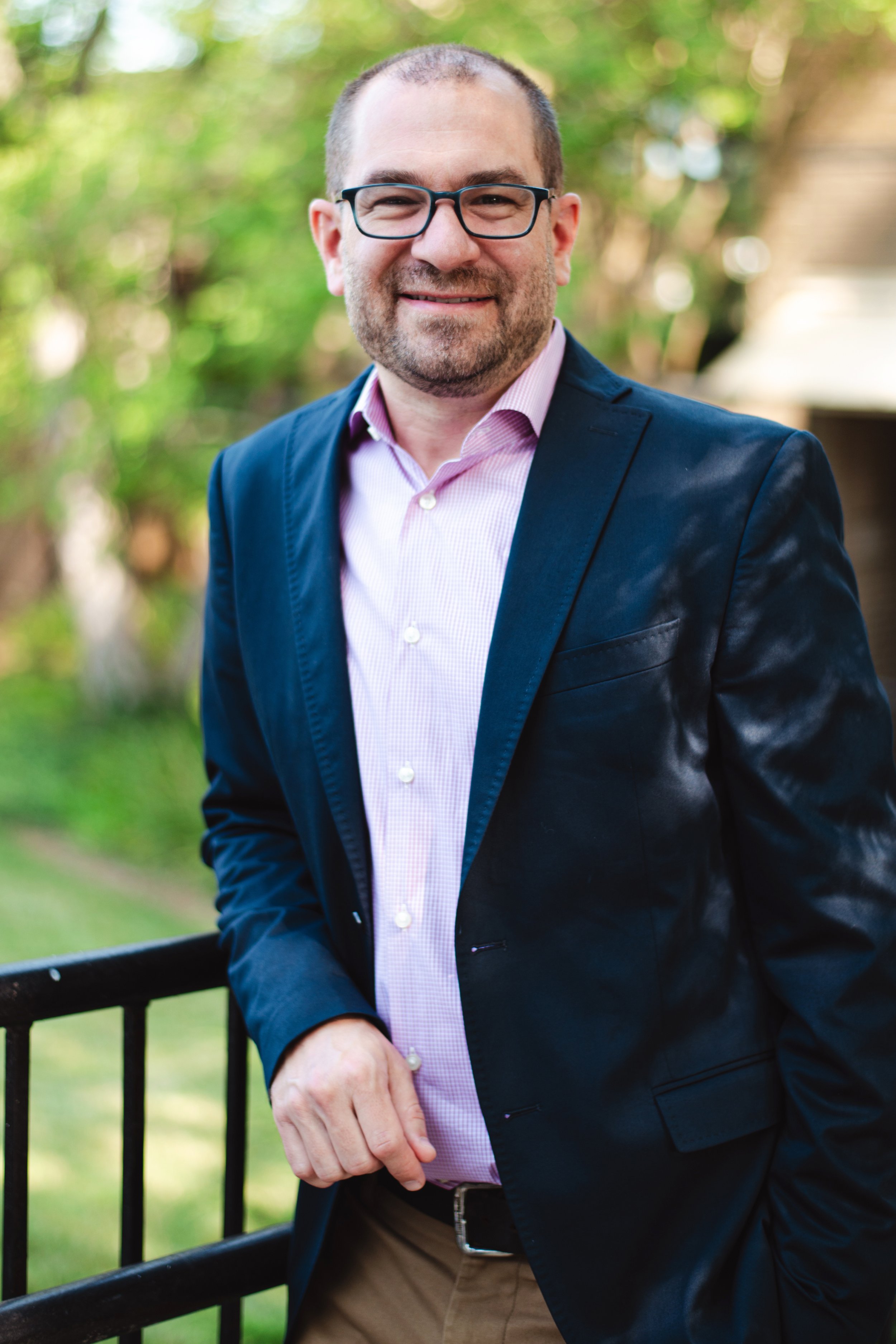 A man with short hair, glasses, and a beard, wearing a navy blazer, pink shirt, and khaki pants, stands outdoors near a black metal railing with a background of green trees and grass