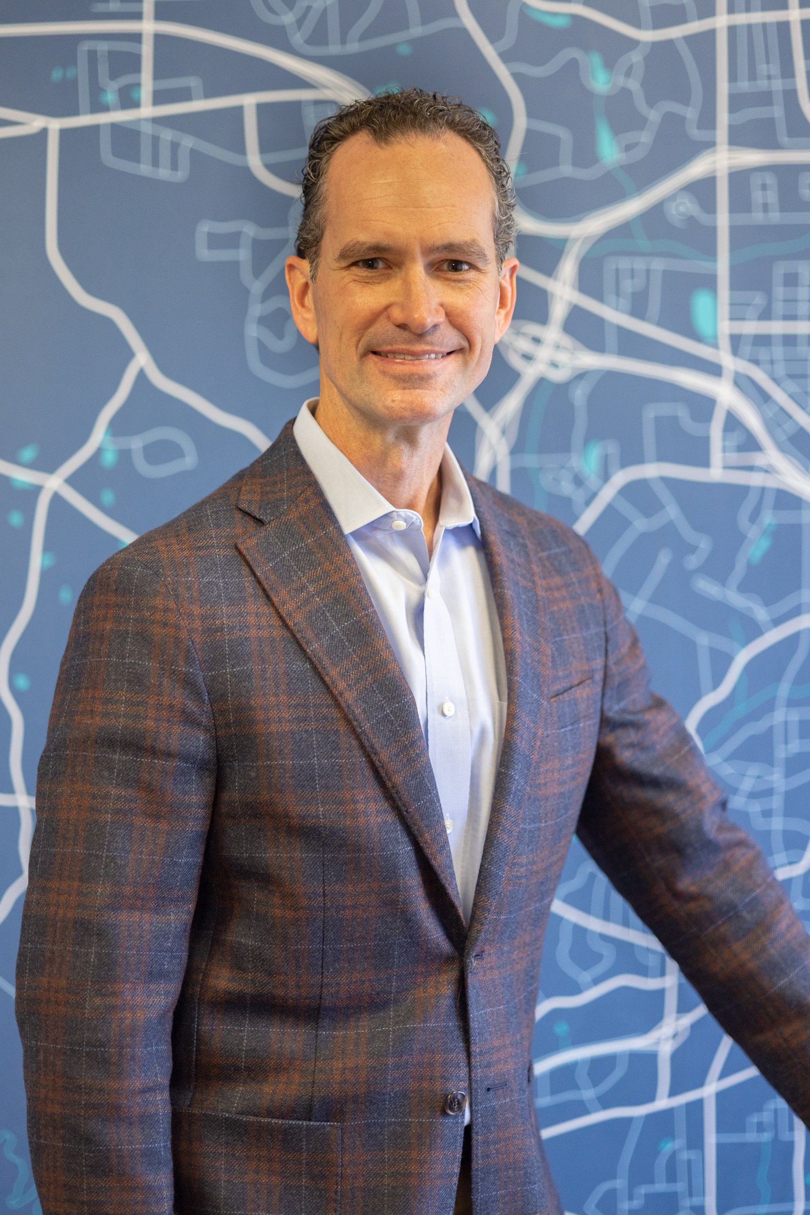 A man in a brown checkered blazer and white shirt standing in front of a blue map or city plan.