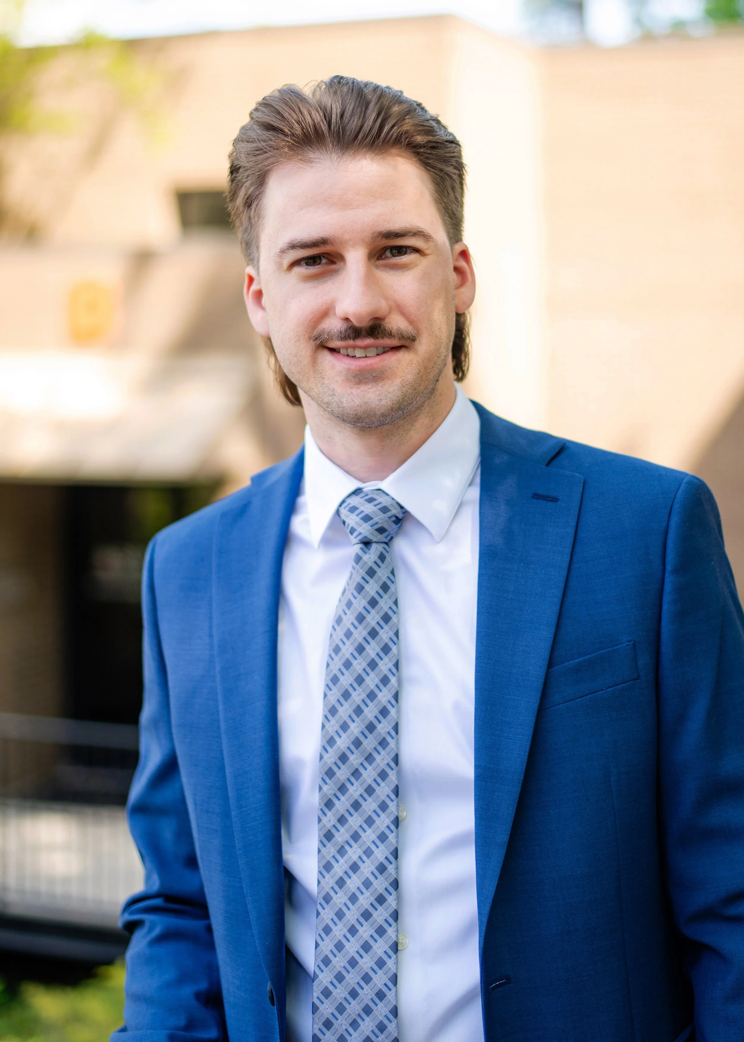 A young man with brown hair and a mustache, wearing a blue suit and a patterned tie, standing outdoors with a blurred background of a building and trees.