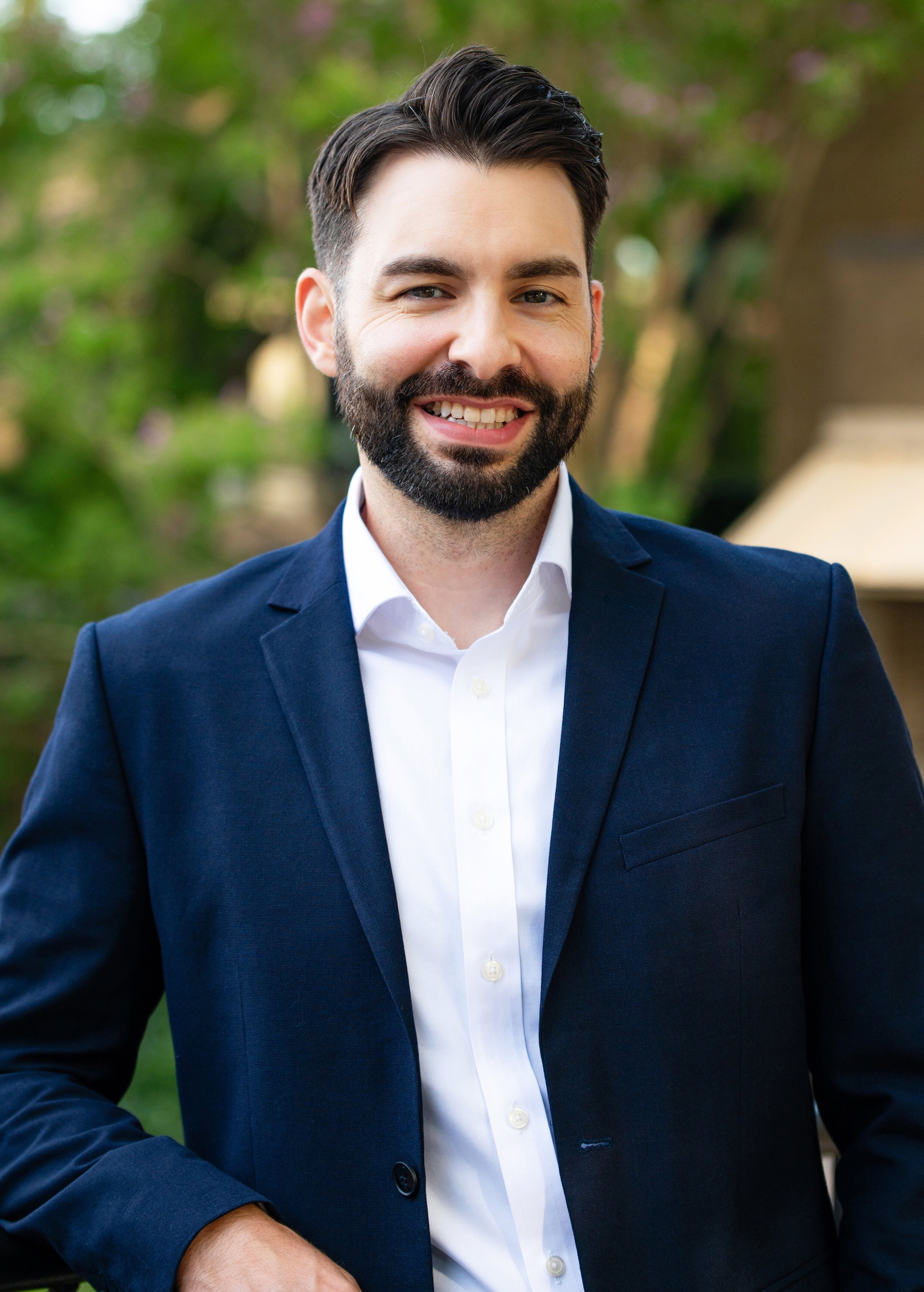 A smiling man with dark hair and a beard, wearing a navy blue blazer and white shirt, standing outdoors with blurred greenery in the background.