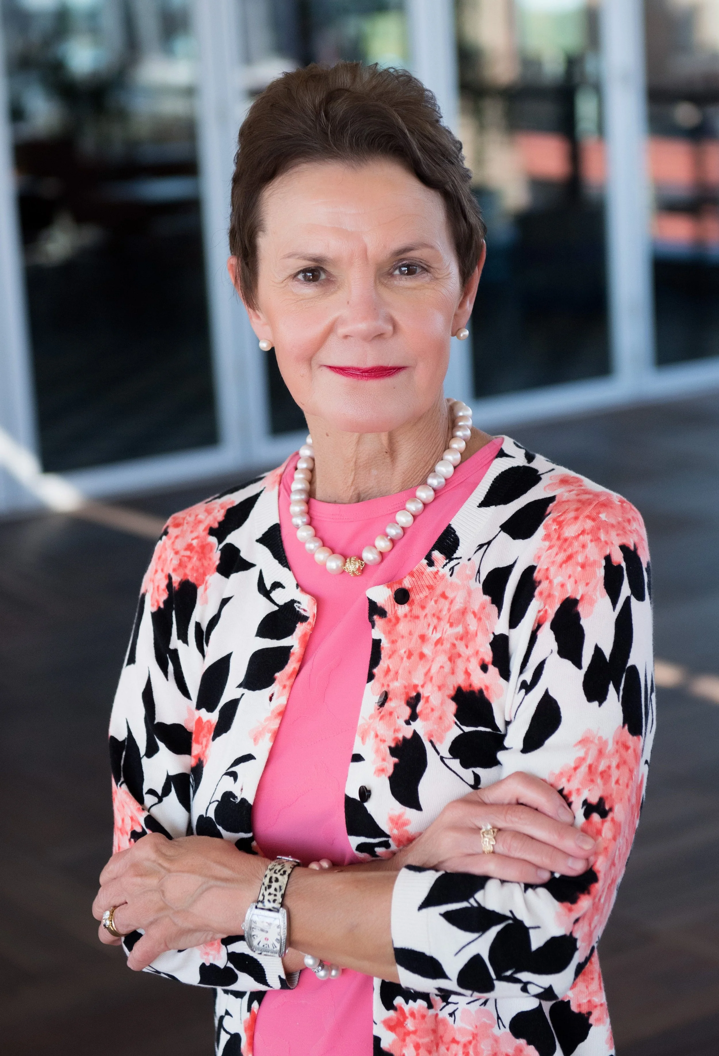 A mature woman with short dark hair, wearing a floral blazer with pink and black patterns, pink blouse, pearl necklace, pearl earrings, watch, and rings, standing with arms crossed in an indoor space with glass sliding doors in the background.