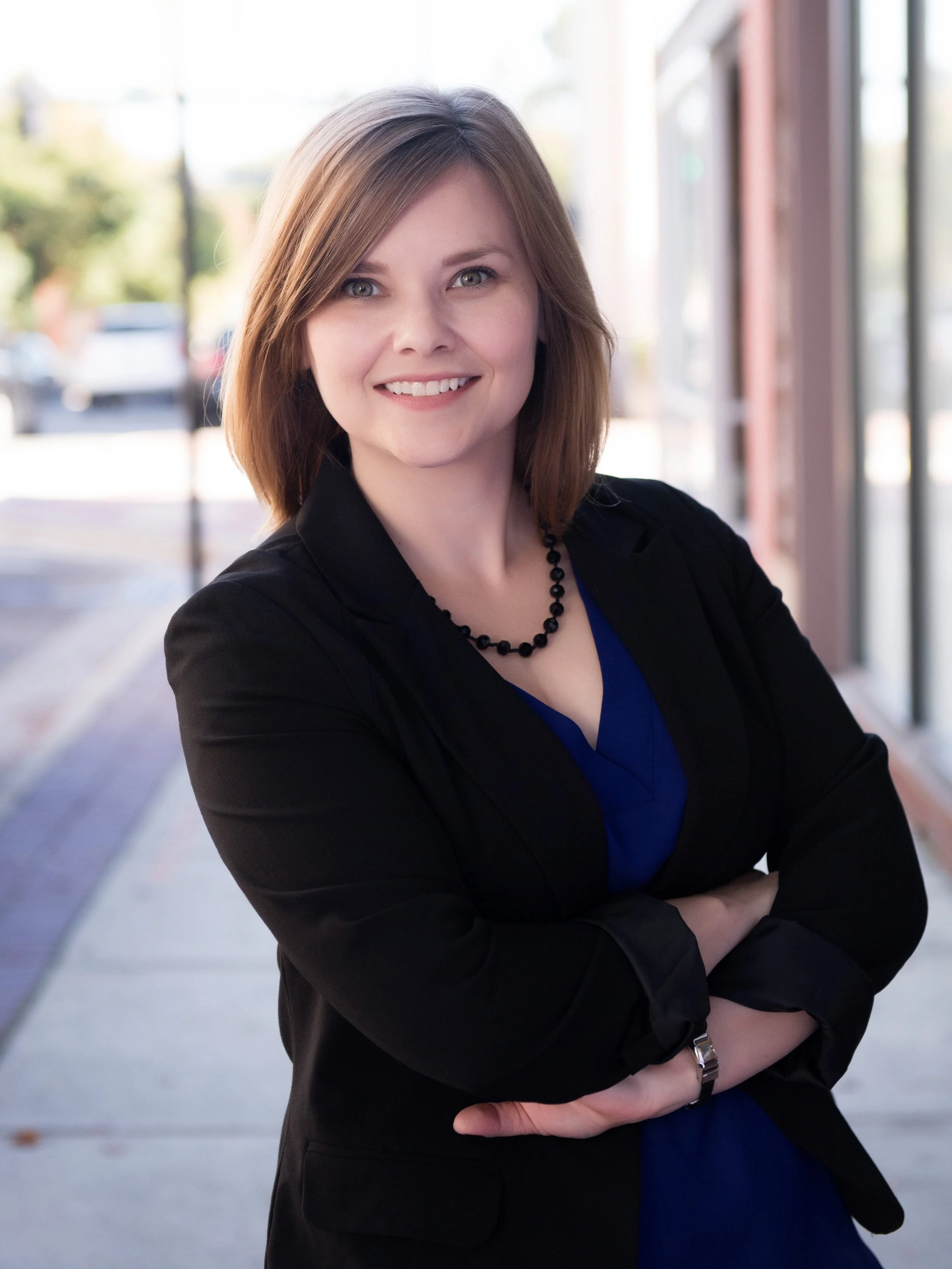 A woman with shoulder-length light brown hair, wearing a black blazer, blue top, black beaded necklace, and a wristwatch, smiling with arms crossed outside on a sidewalk with blurred trees and buildings in the background.