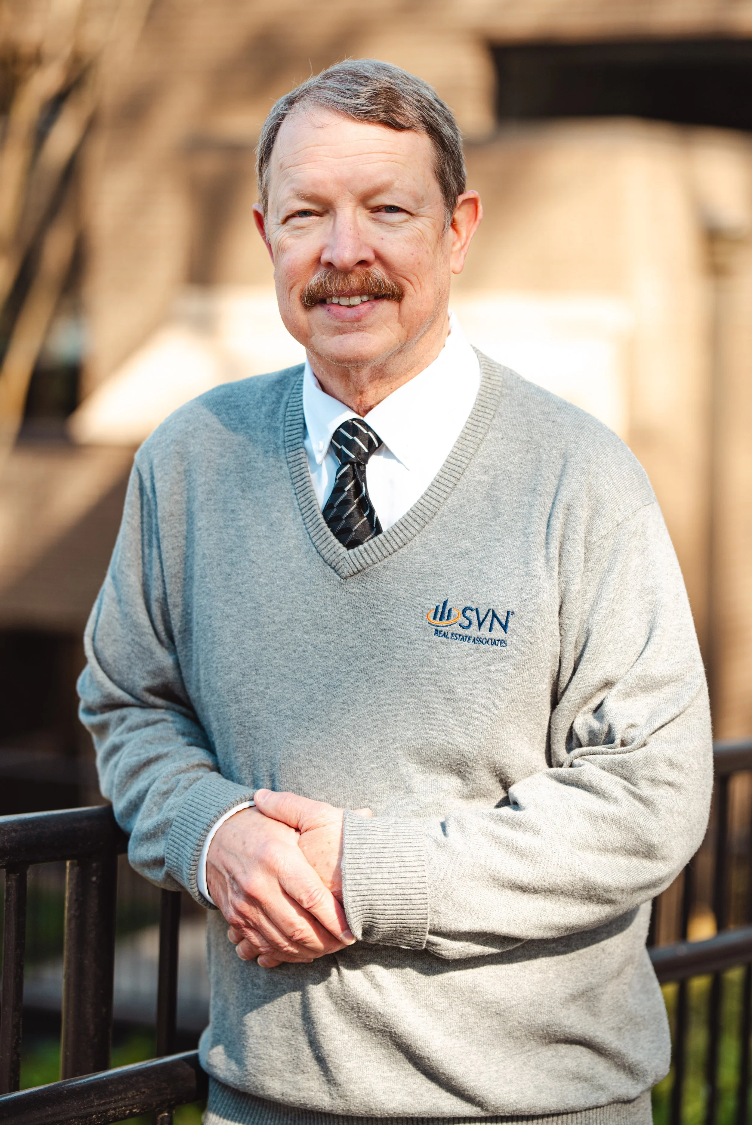 A middle-aged man with a mustache and light brown hair, wearing a gray sweater with a logo, a white dress shirt, and a black patterned tie, standing outdoors in front of a blurred background of a building, smiling and clasping his hands in front of him.