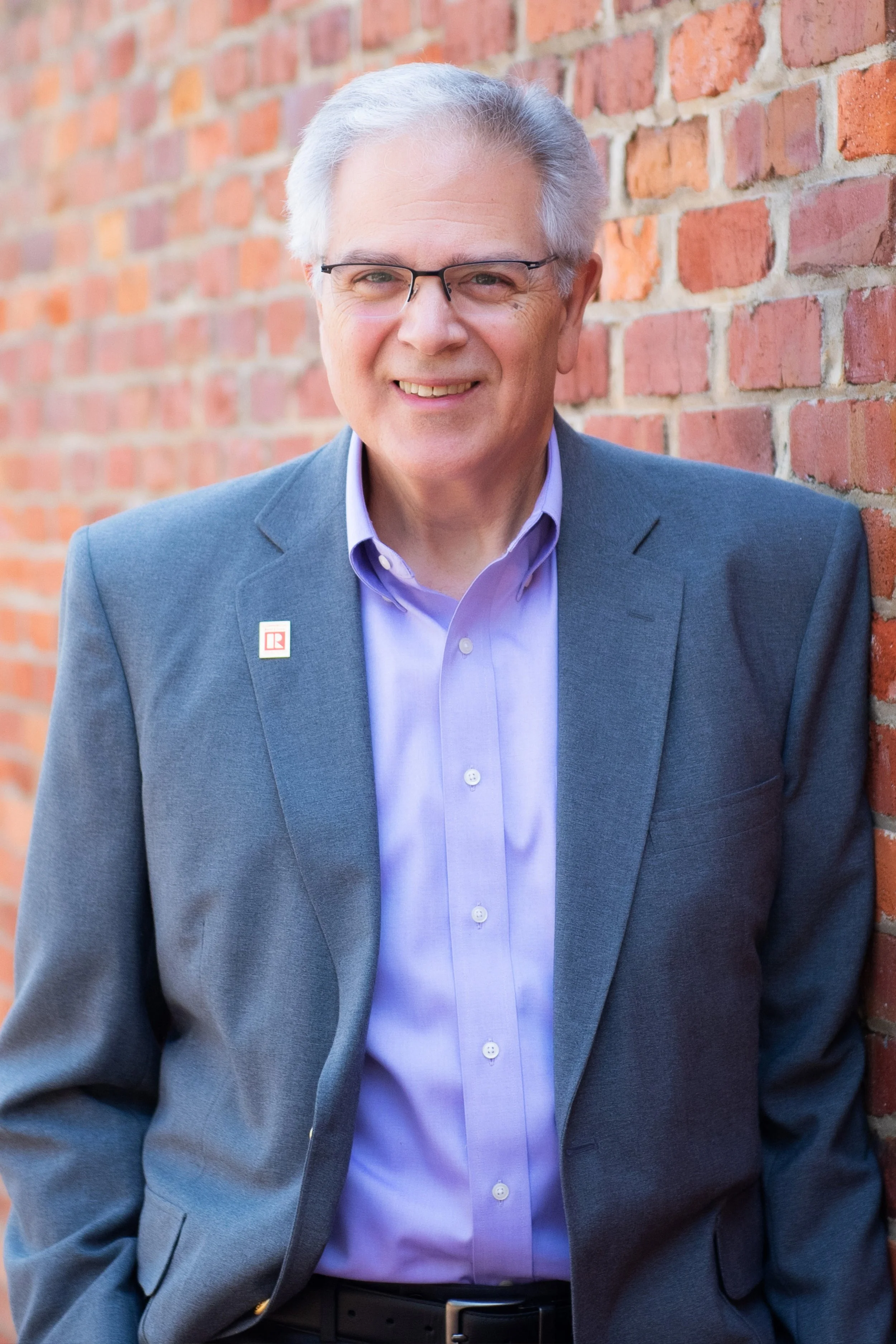 A smiling older man with gray hair and glasses, wearing a gray suit jacket and a light purple shirt, leaning against a red brick wall outdoors.