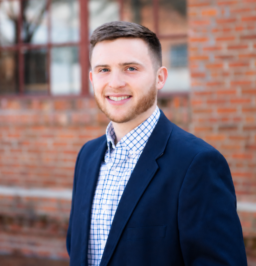 A young man with short brown hair, blue eyes, and a beard, smiling, wearing a navy blazer and a blue and white checkered shirt, standing outdoors in front of a brick building with windows.