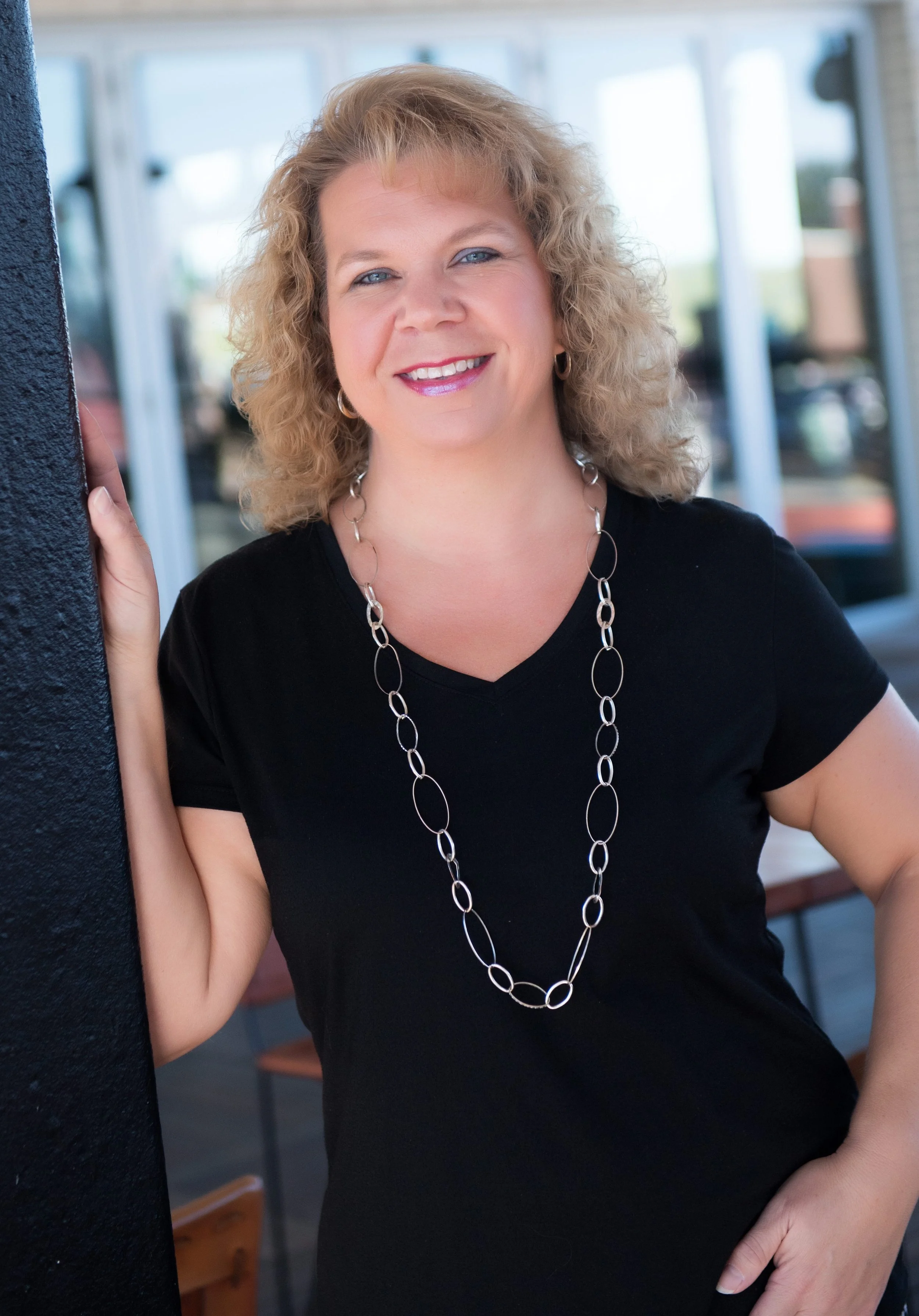 A woman with curly blonde hair and blue eyes smiling, wearing a black V-neck shirt and a long silver chain necklace, standing outdoors near a building with glass windows.