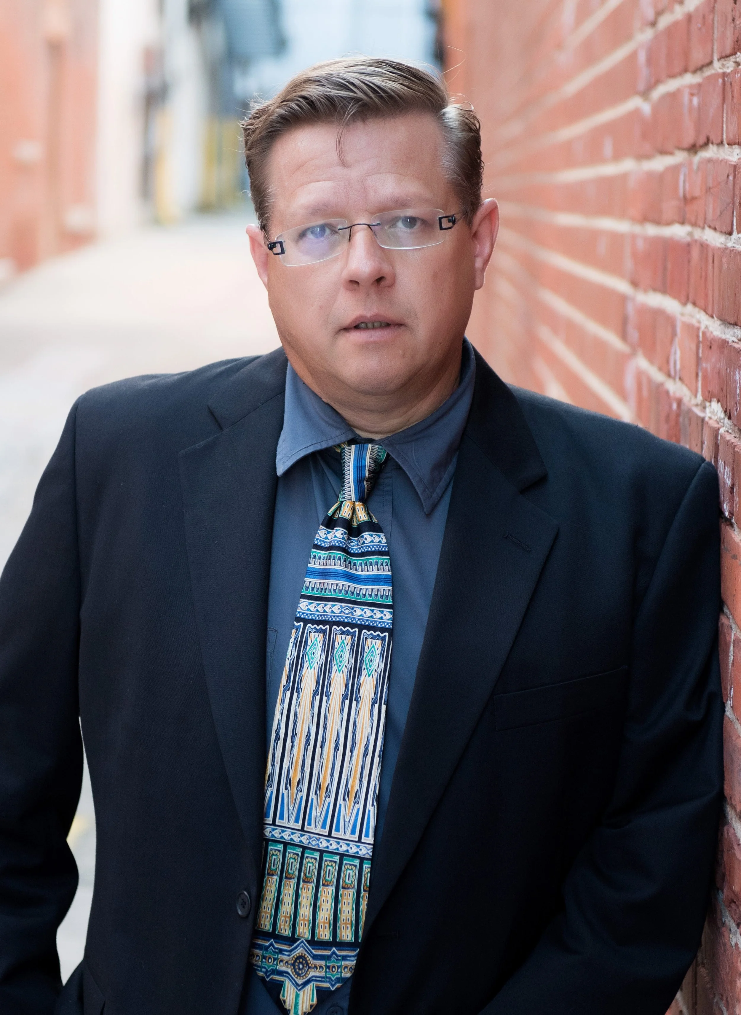 A middle-aged man with glasses and short hair, wearing a dark suit, blue shirt, and a colorful patterned tie, leaning against a brick wall in an alleyway.
