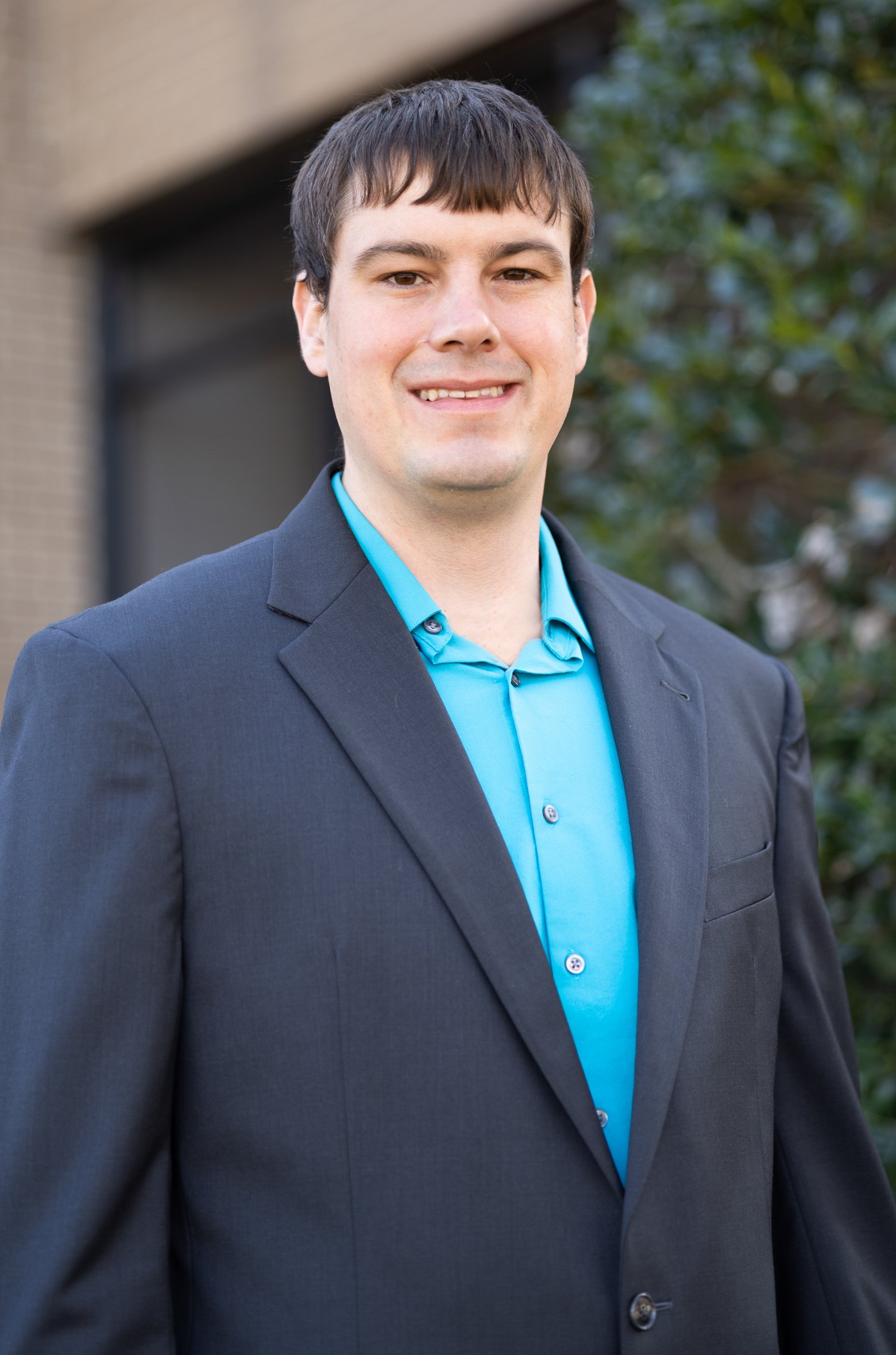 A young man with short brown hair wearing a dark gray suit and a light blue collared shirt standing outdoors near a building and greenery.