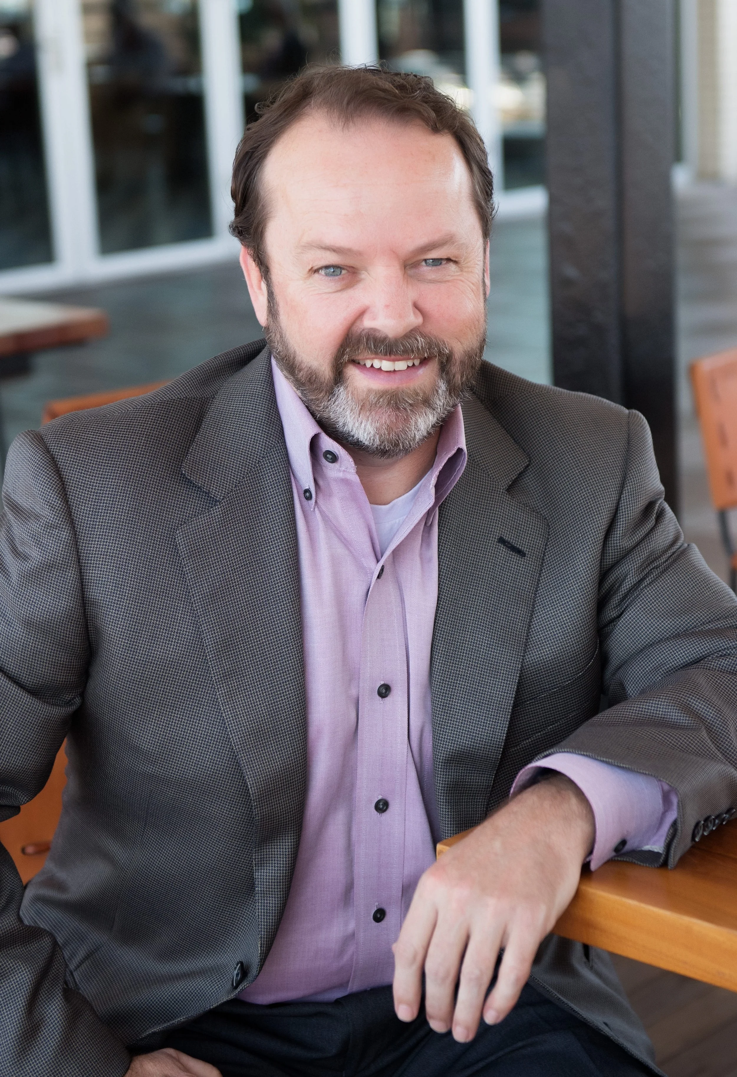 A man with a beard and mustache, wearing a grey blazer and a lavender dress shirt, is sitting at a wooden table in an outdoor setting with glass windows and chairs in the background, smiling at the camera.