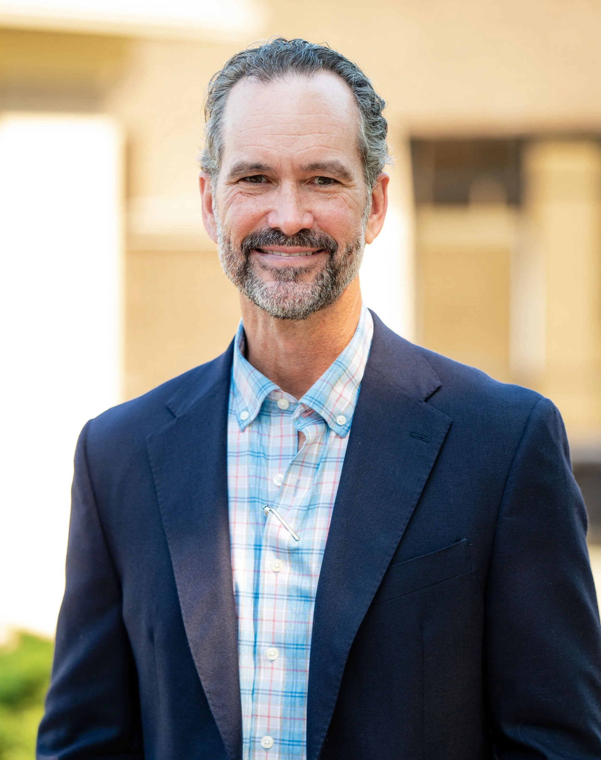 A man wearing a brown plaid blazer and white shirt standing against a blue wall with a map background.
