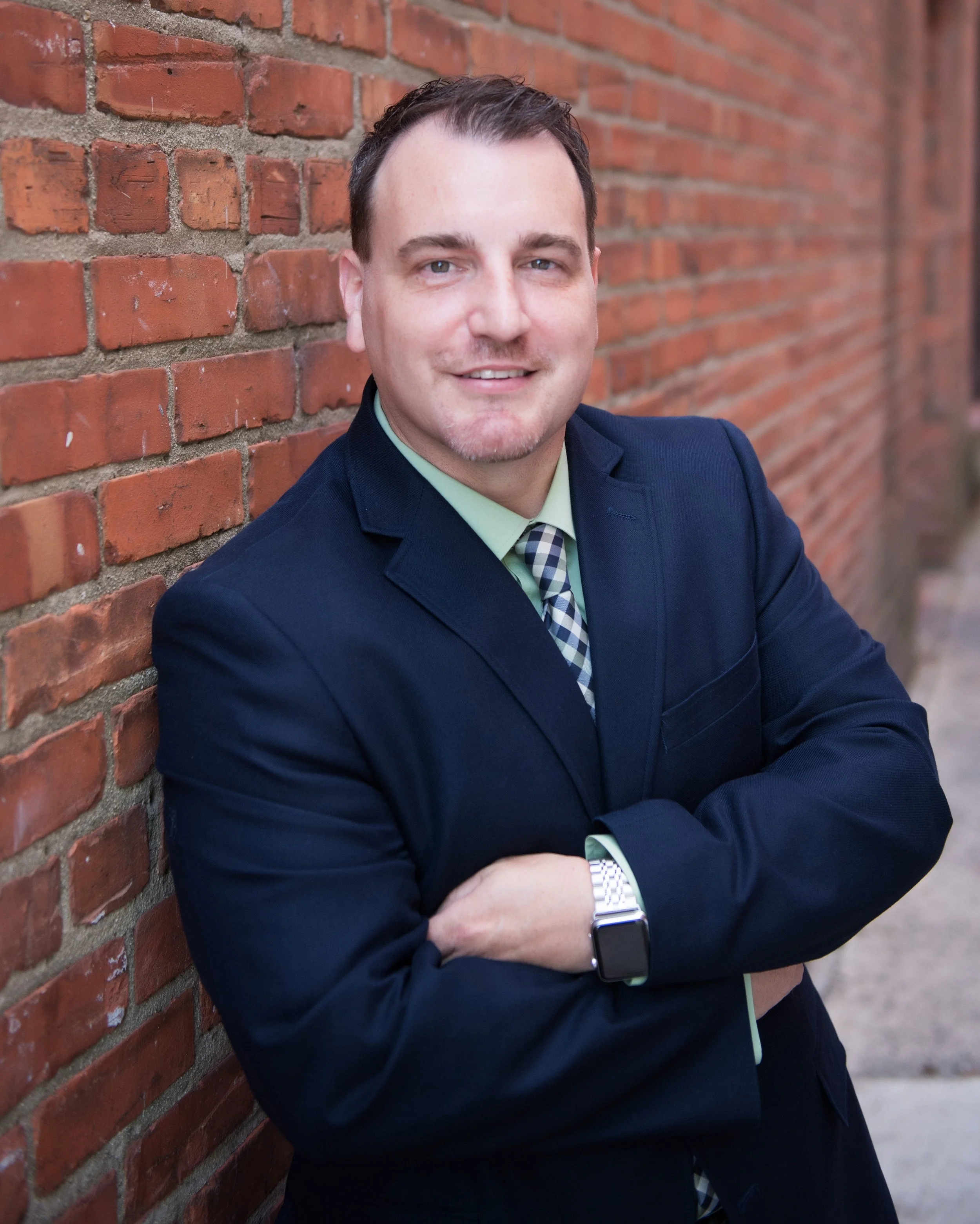 A man in a navy suit with a green shirt and plaid tie standing against a brick wall with arms crossed, smiling.