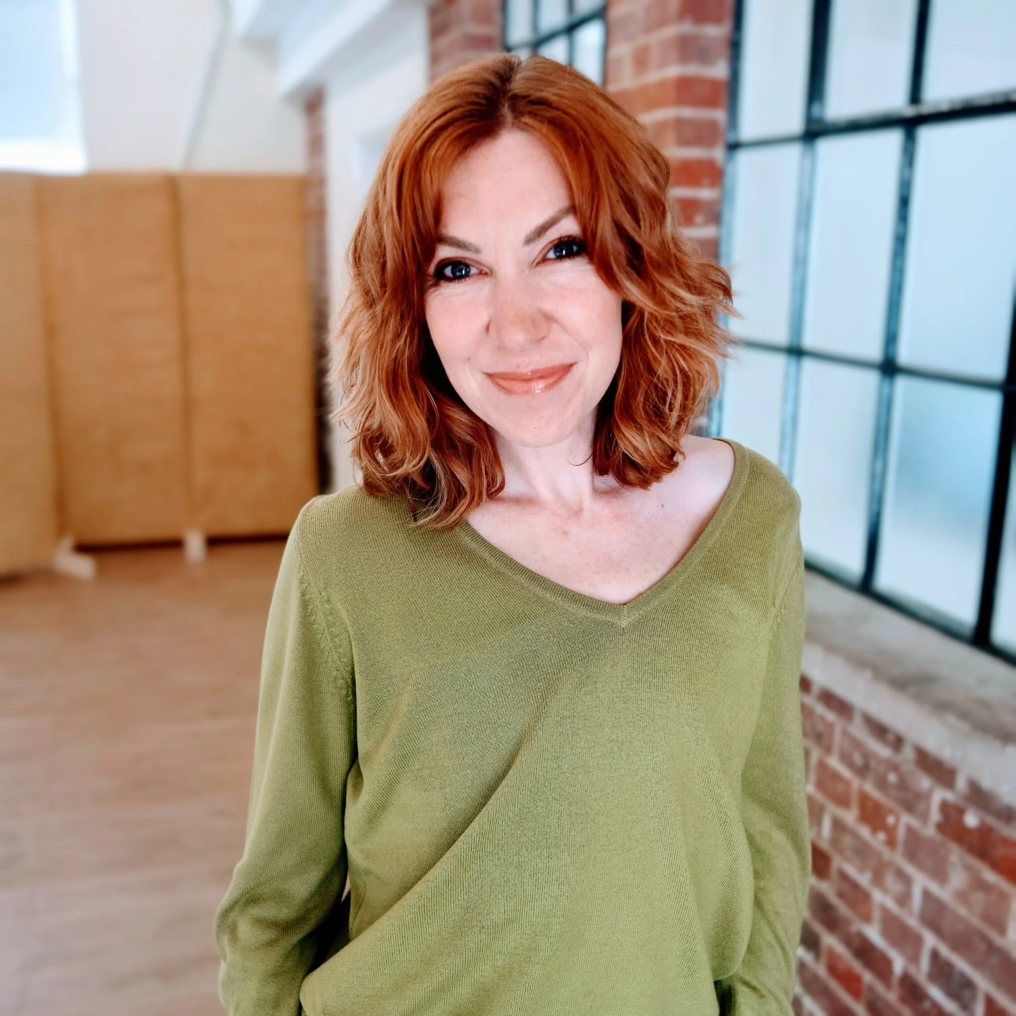 Close-up of a woman with red hair and blue eyes smiling, wearing a green shirt, in an indoor setting with brick walls and large windows. Samantha Vale - Founder of Improving People.