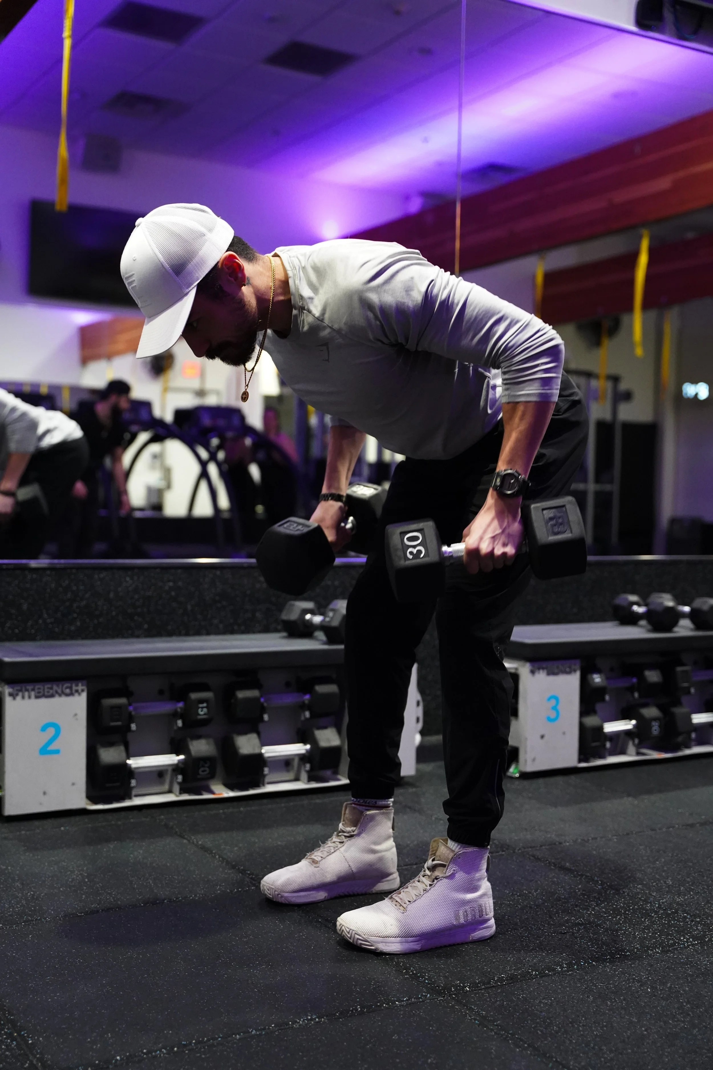 A man lifting dumbbells in a gym, bending forward with a mirror behind him showing more gym equipment and people working out.