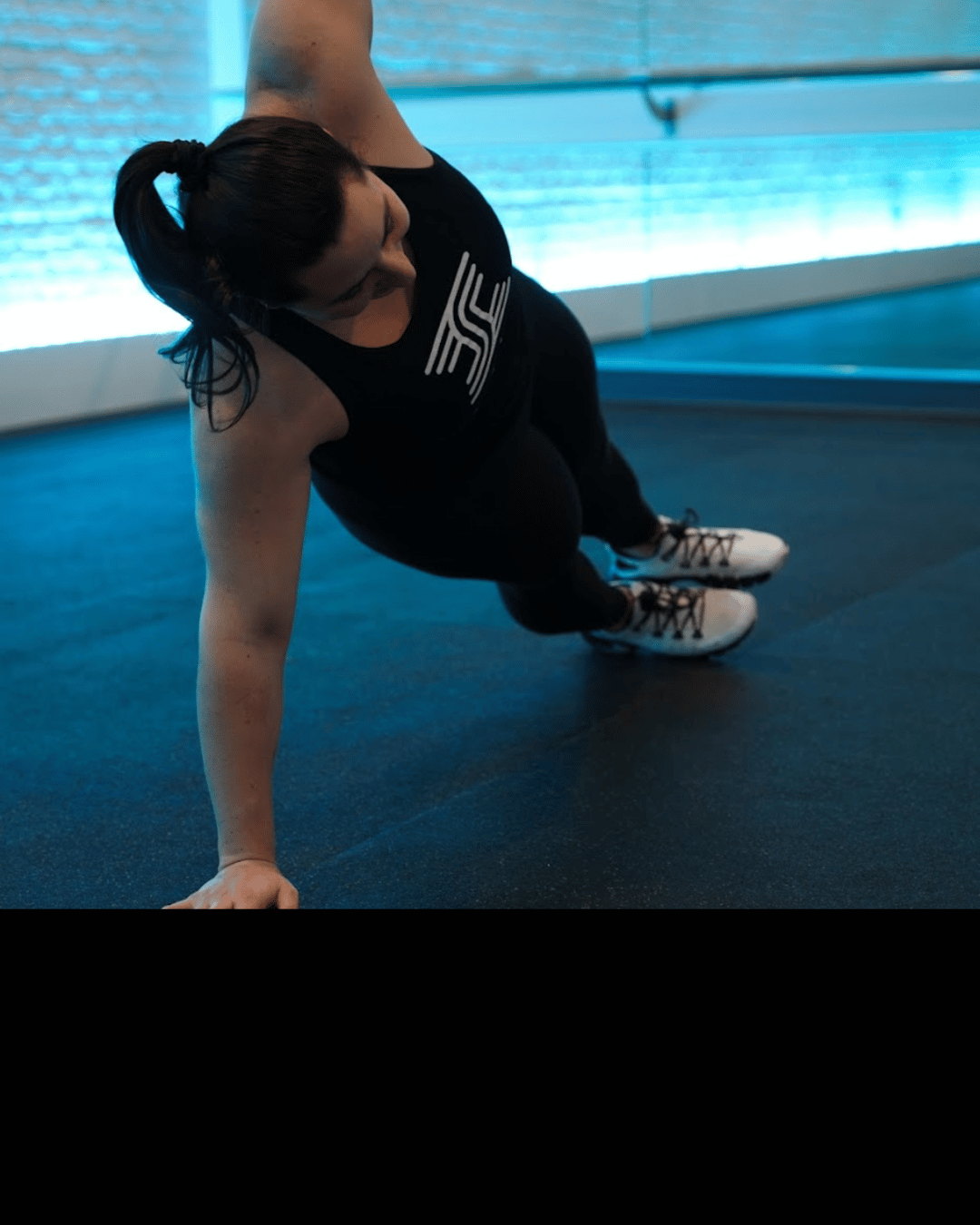 A woman in black athletic clothing and white sneakers doing a push-up in a gym with blue lighting.