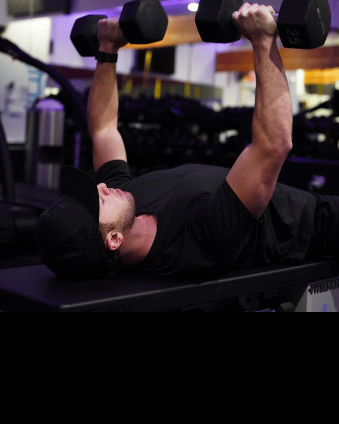 A man lying on a workout bench performing a dumbbell bench press in a gym, wearing a black cap, black shirt, and holding two dumbbells.