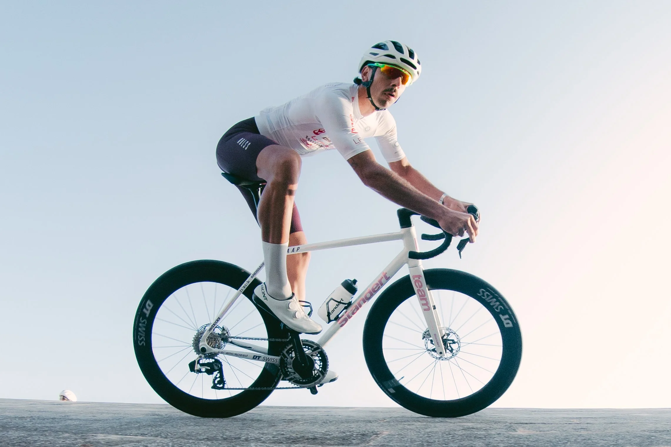 A cyclist riding a standert kreissäge rs white road bike on a beach with the ocean in the background, wearing a white helmet, sunglasses, and athletic clothing.