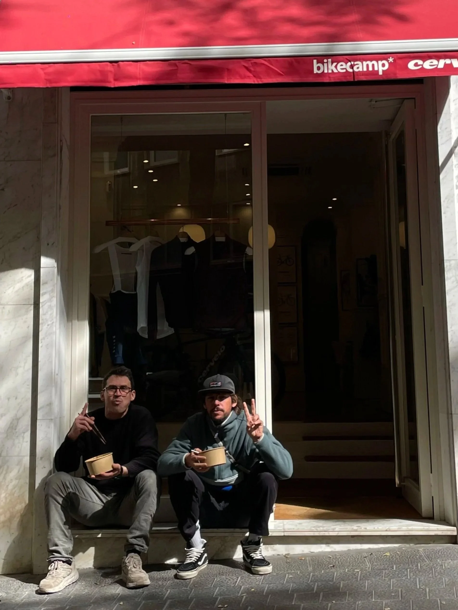 Two owner sitting on the sidewalk outside a storefront, holding bowls and chopsticks, making peace signs and posing for the camera. The storefront has large glass windows and a red awning with visible branding.