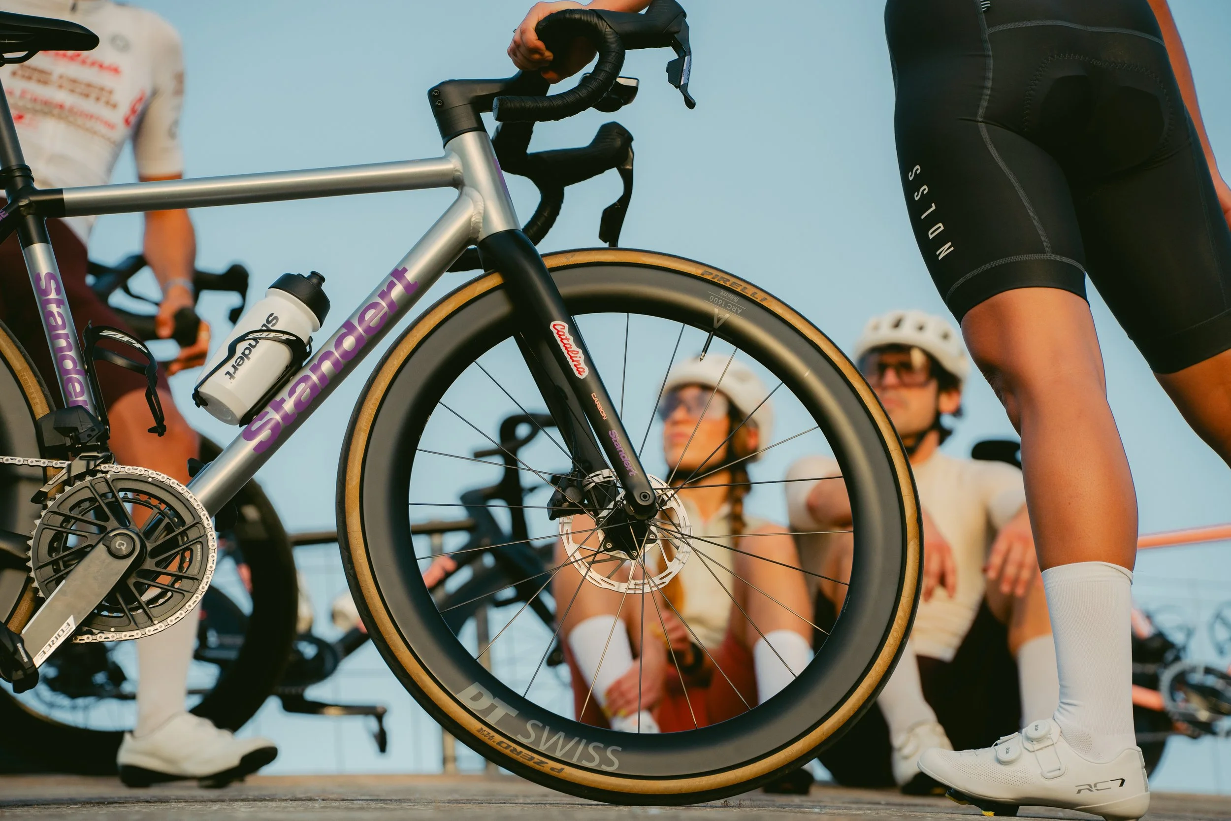 Close-up of a cyclist's hand gripping the handlebars of a road bike, with several people in the background wearing helmets and athletic attire outdoors during daylight.