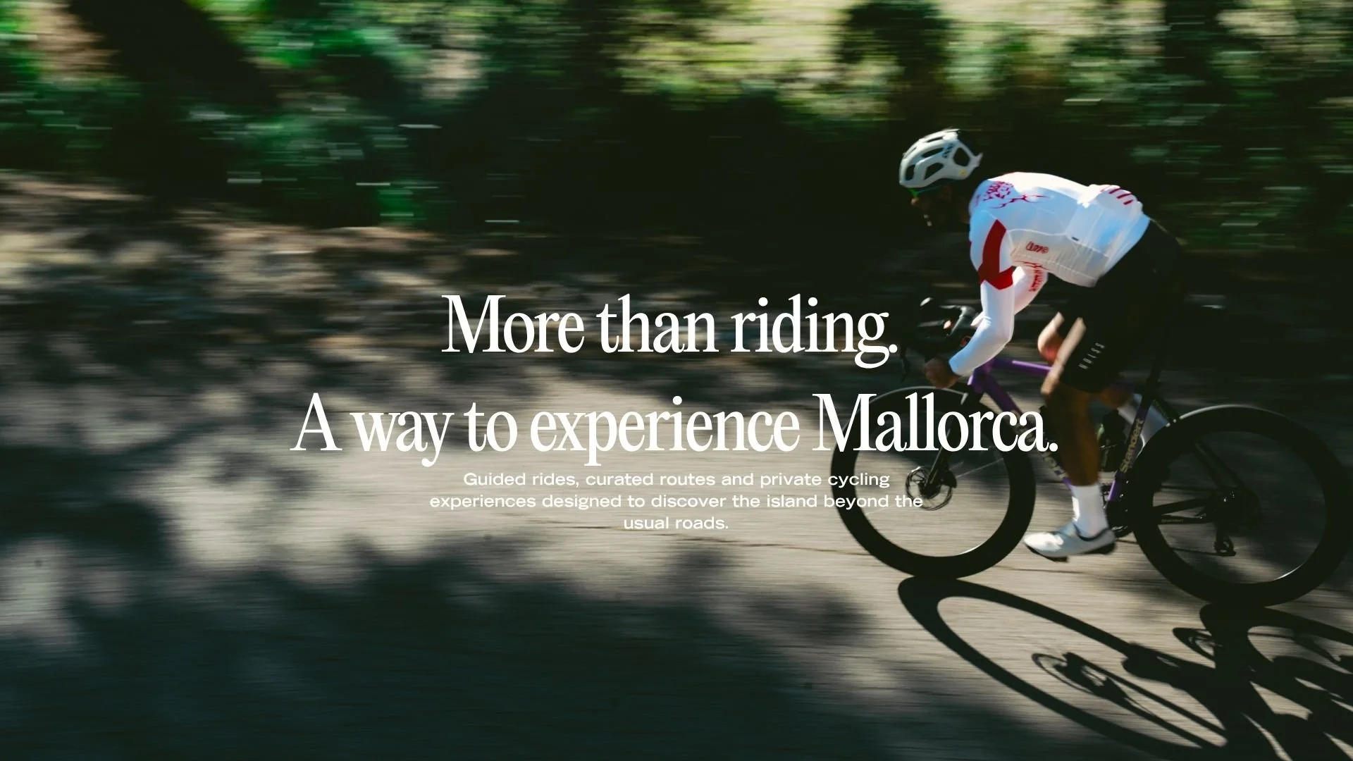 A person riding a bicycle in motion through a forested area with blurred trees in the background, wearing a white helmet and sportswear. Overlaid text says, 'More than riding. A way to experience Mallorca. Guided rides, curated routes and private cycling experiences designed to discover the island beyond the usual roads.'