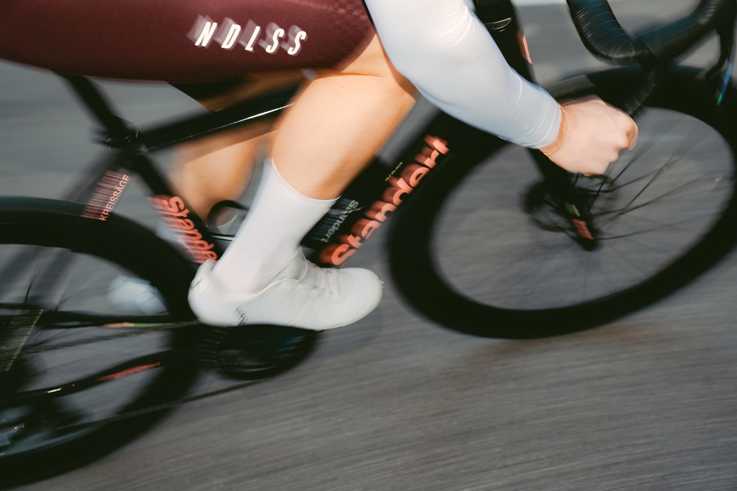 Close-up of a person riding a standert kreissäge black bike, wearing NDLSS white socks, BONT white shoes, and a NDLSS dark red bib with white text, with motion blur indicating speed.