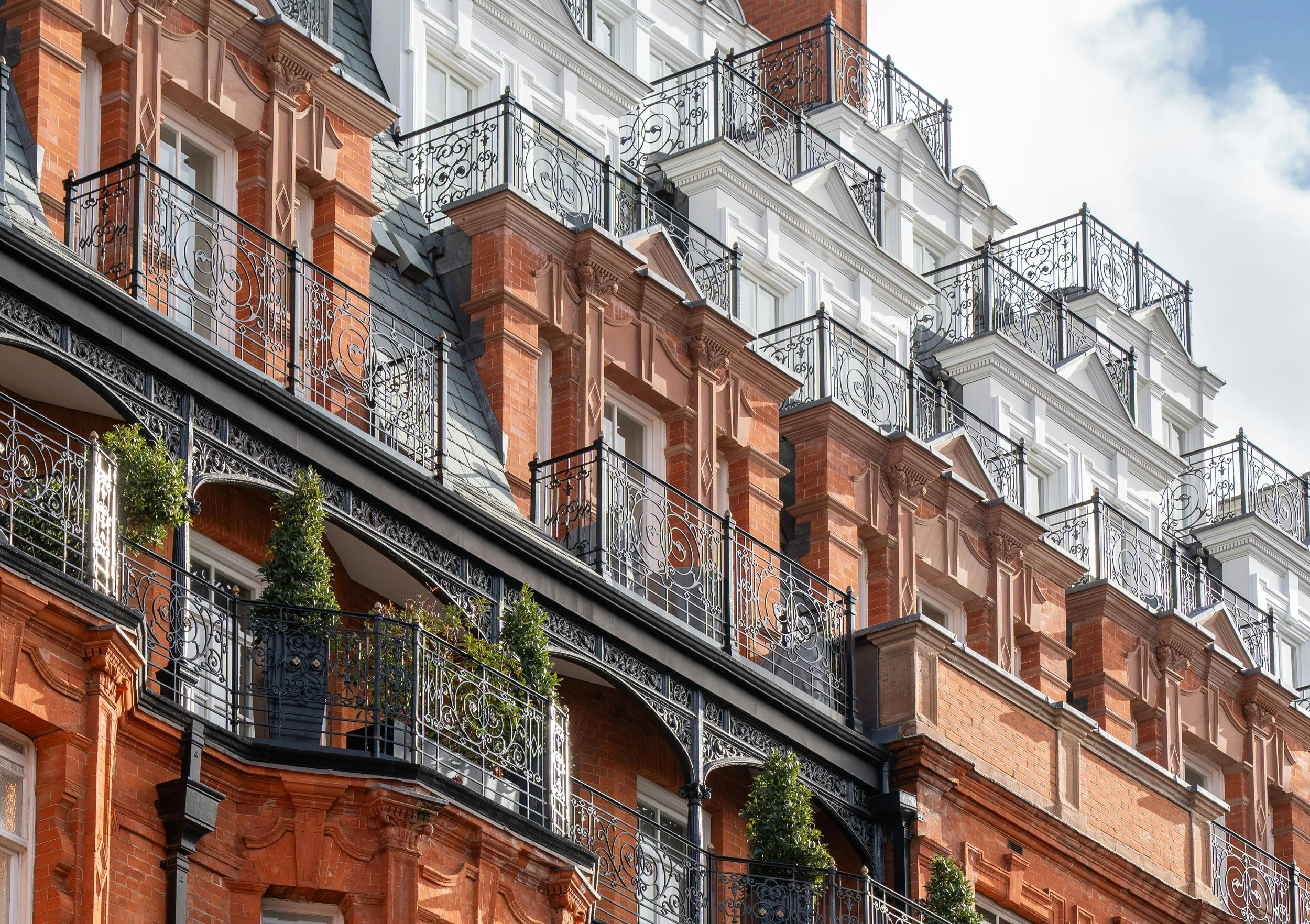 Close-up of a multi-story brick building with ornate black wrought-iron balconies, some with potted plants. The building features decorative architectural details and white window frames, with a partly cloudy sky in the background.