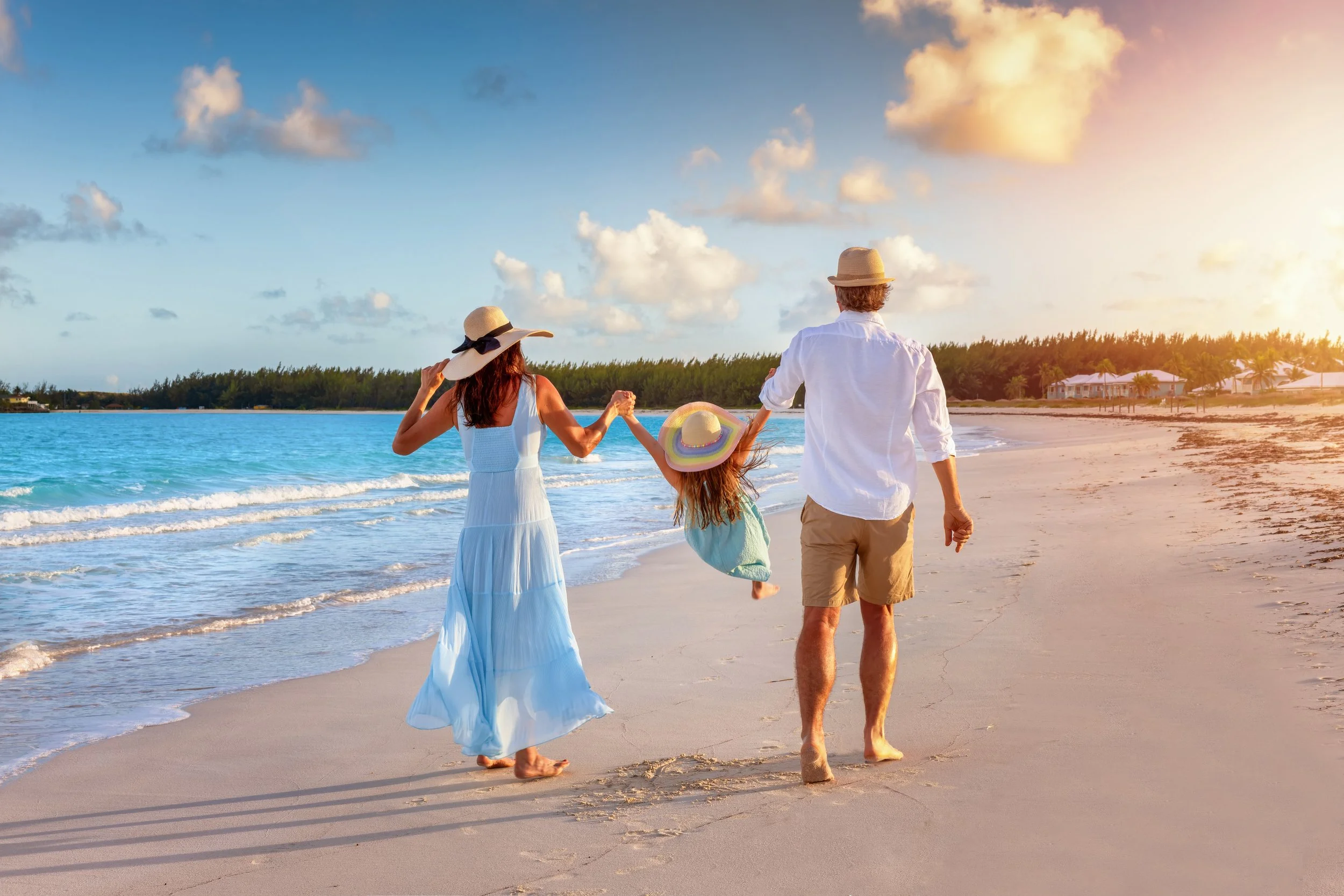 A family of three walking on a sandy beach near the ocean during sunset. The woman is holding the child's hand, and they are all wearing sun hats. The child is swinging by the adults, and the sky is partly cloudy.