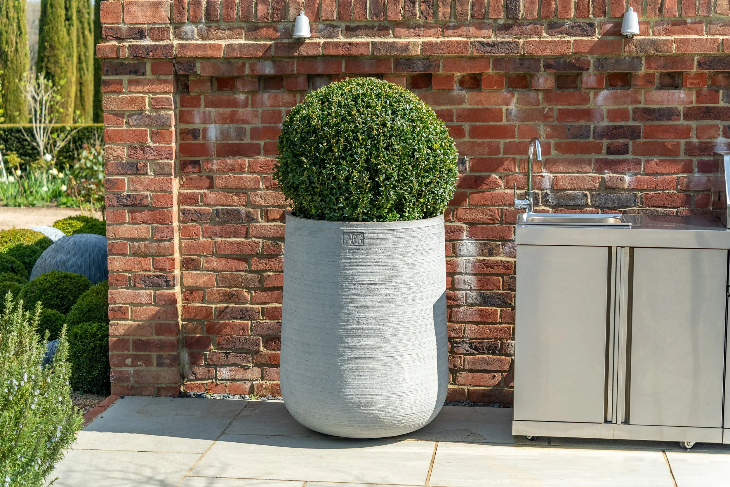 Large white planter with a lush green shrub against a red brick wall next to a stainless steel outdoor sink cabinet.