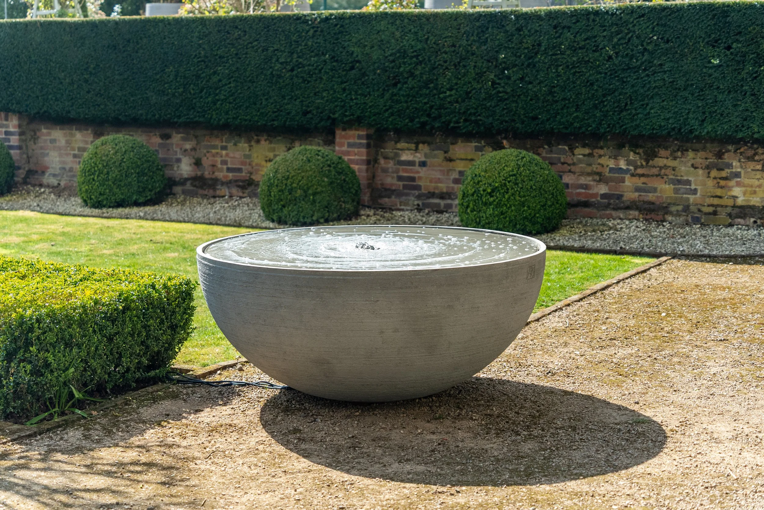Stone bowl fountain with water, outdoor garden with trimmed bushes, brick wall, and lawn.
