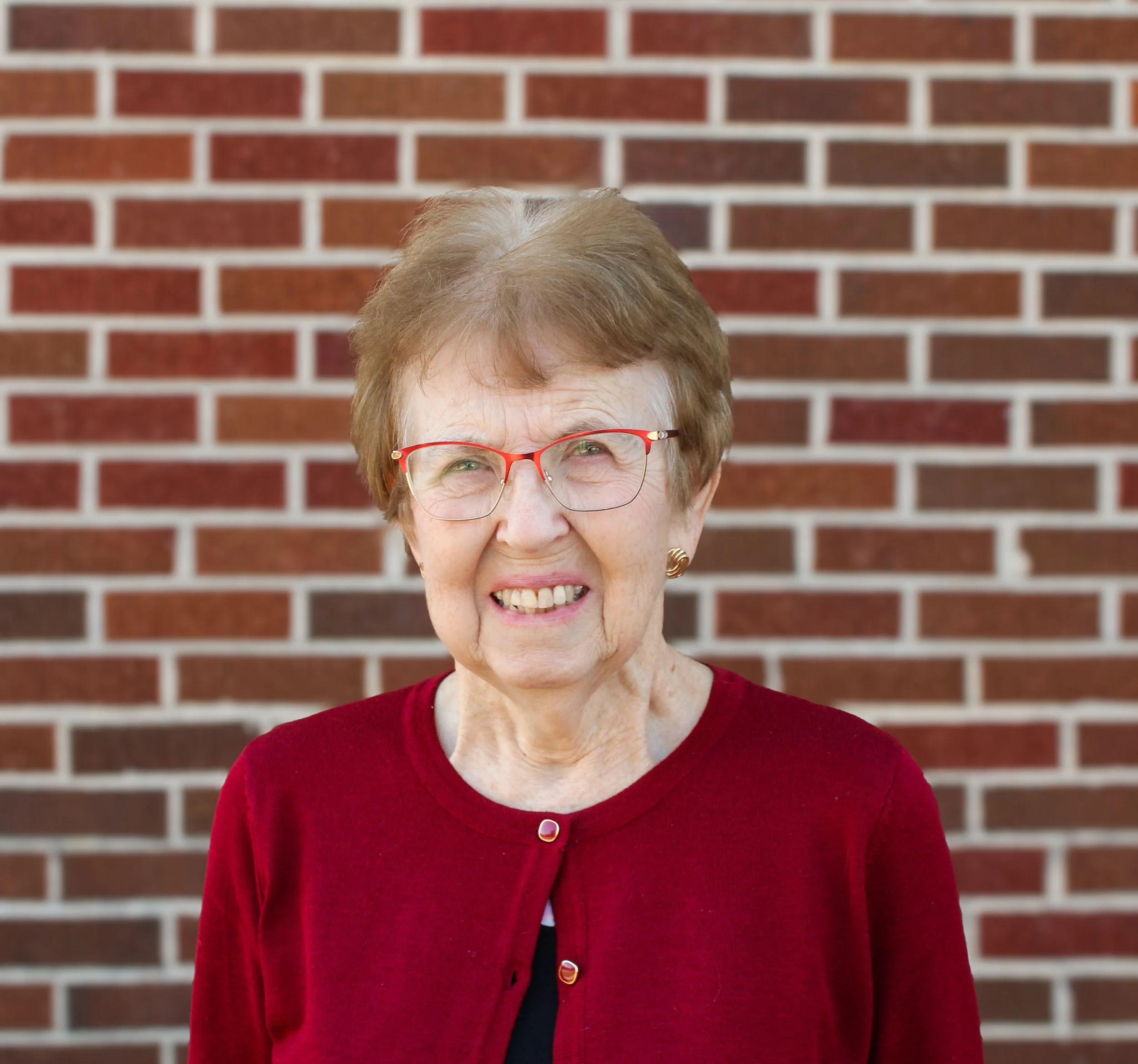 An elderly woman with short light brown hair, wearing glasses and a red cardigan, sitting at a piano and playing it.