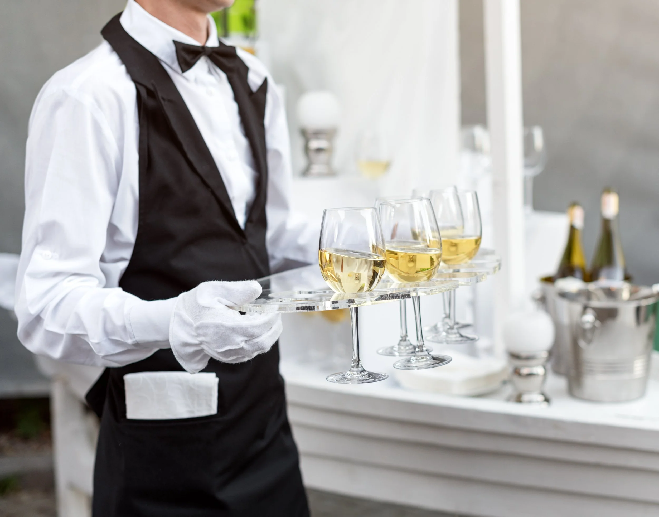 Person dressed as a waiter holding a tray with four glasses of white wine, wearing a tuxedo with a bow tie and gloves, standing in front of a bar with bottles and glasses.