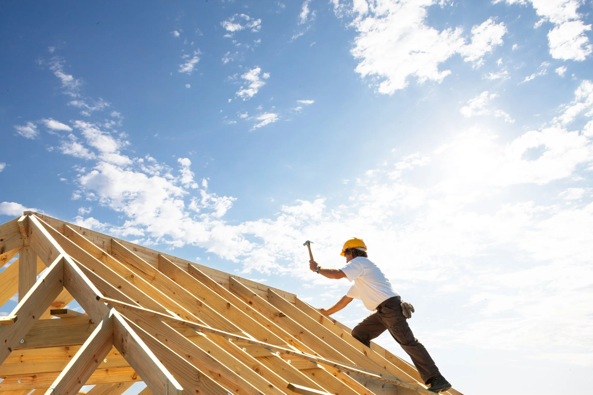 A construction worker wearing a yellow helmet and white shirt working on a wooden roof frame with a hammer against a blue sky with white clouds.