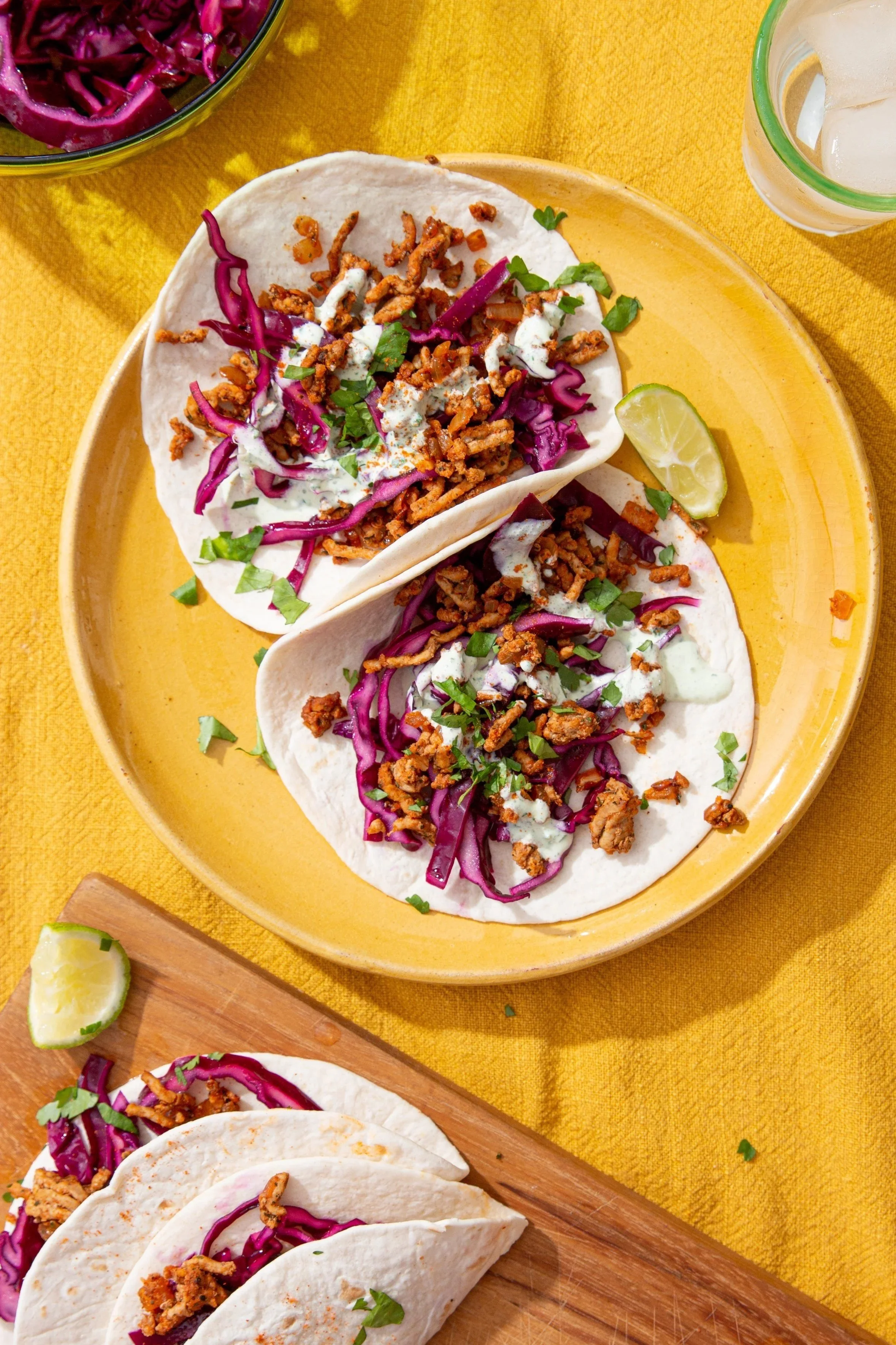 Two soft tacos filled with shredded purple cabbage, cooked meat, and topped with sauce and cilantro, served on a yellow plate with lime wedges. Additional tacos and a bowl of shredded purple cabbage are visible in the background on a yellow tableclot