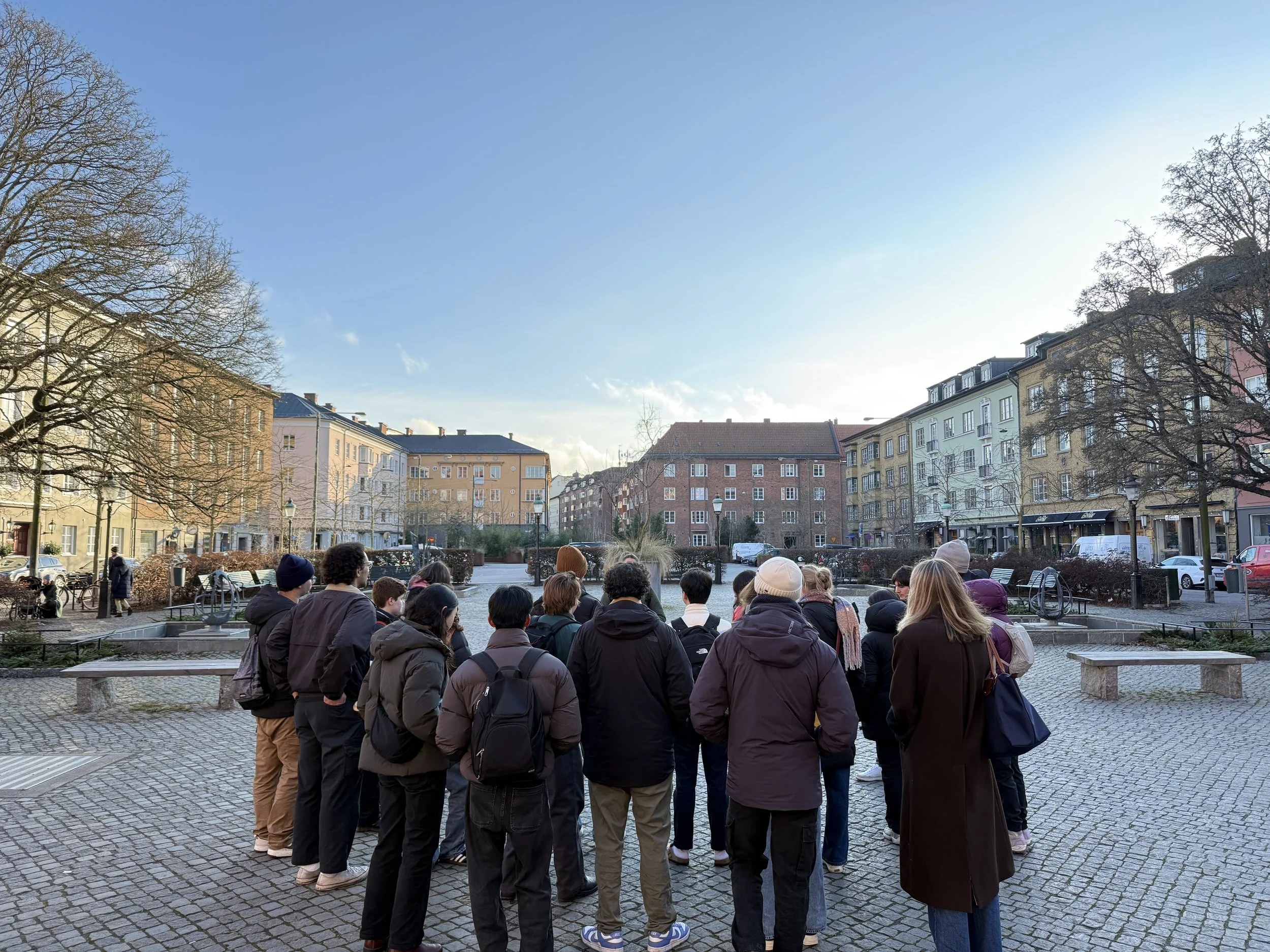 Group of people gathered outdoors in a paved square, facing away, with residential buildings and trees in the background.