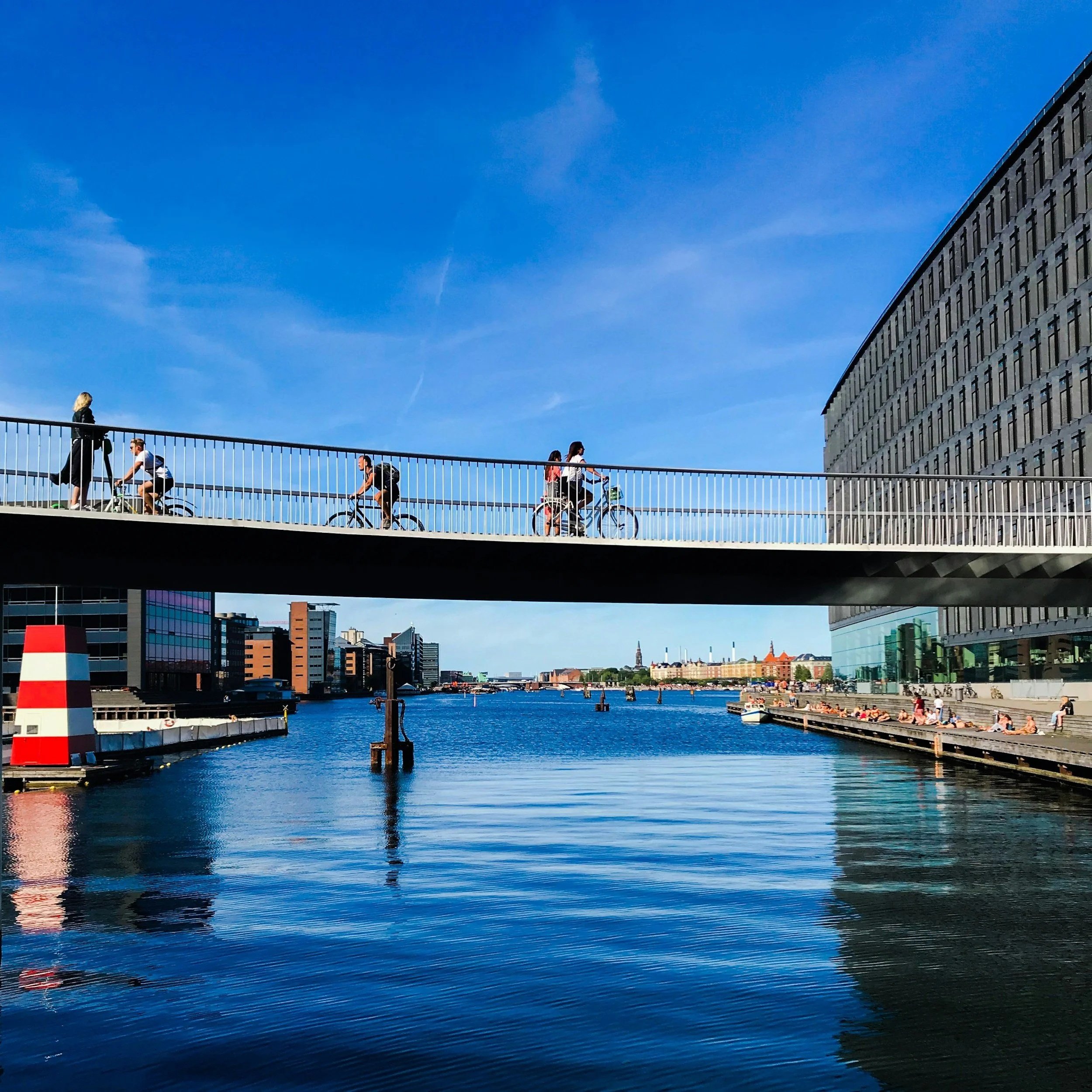 People riding bikes across a modern pedestrian bridge over a river in an urban area with buildings and clear blue sky.
