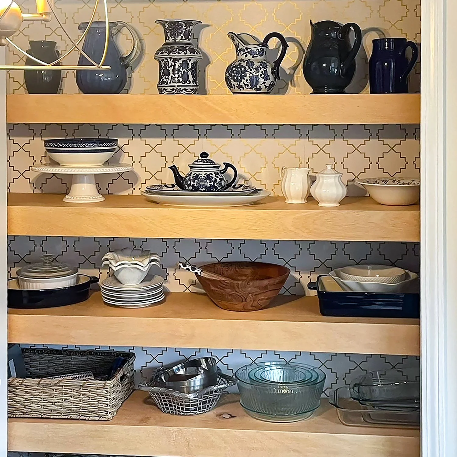 Open pantry shelves with various dishes and kitchen items, including blue and white ceramic pitchers, teapots, a wooden bowl, and glass bowls, with patterned backsplash.