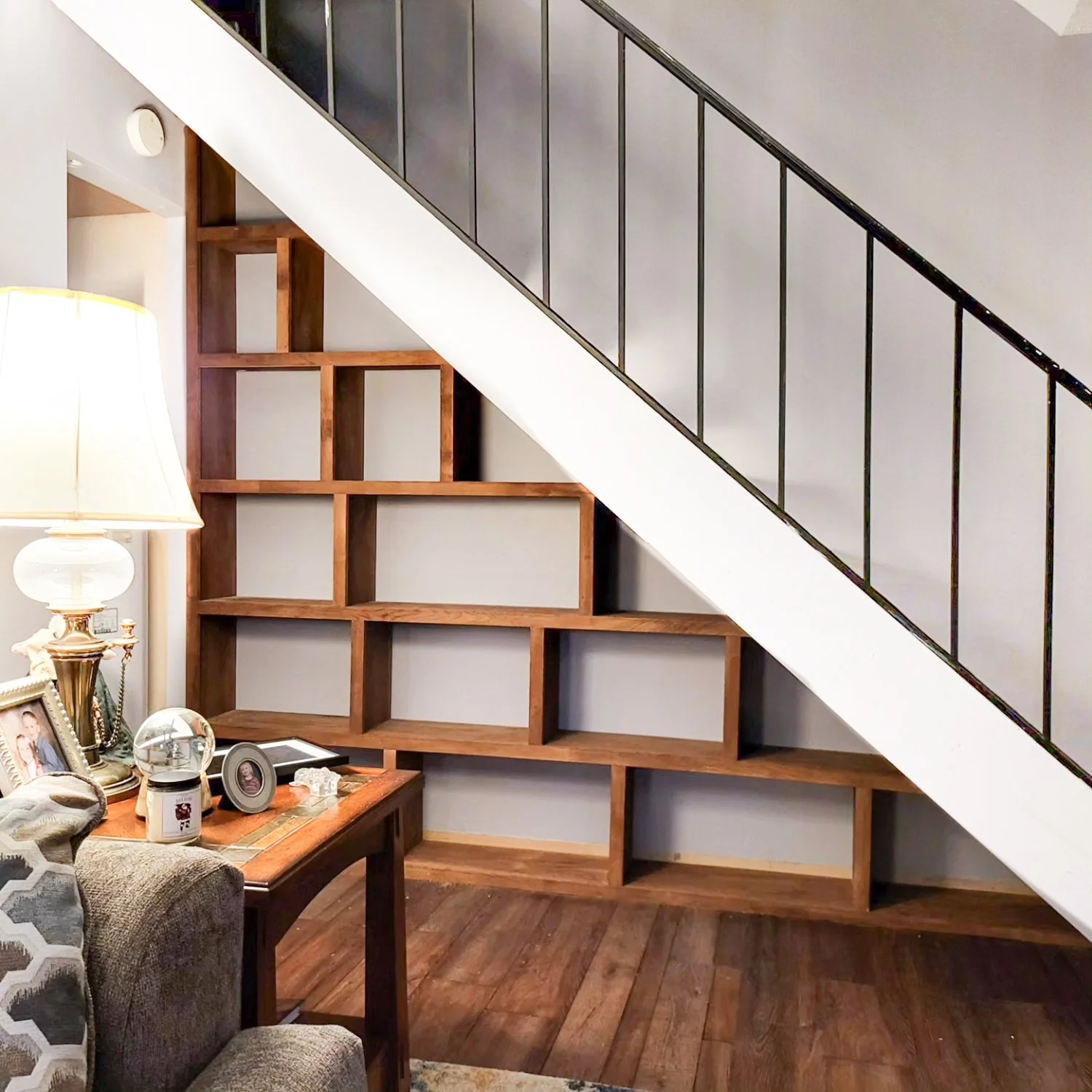 Living room with a staircase featuring built-in wooden shelves, a gray sofa, and a wooden side table with a lamp and decorative items.