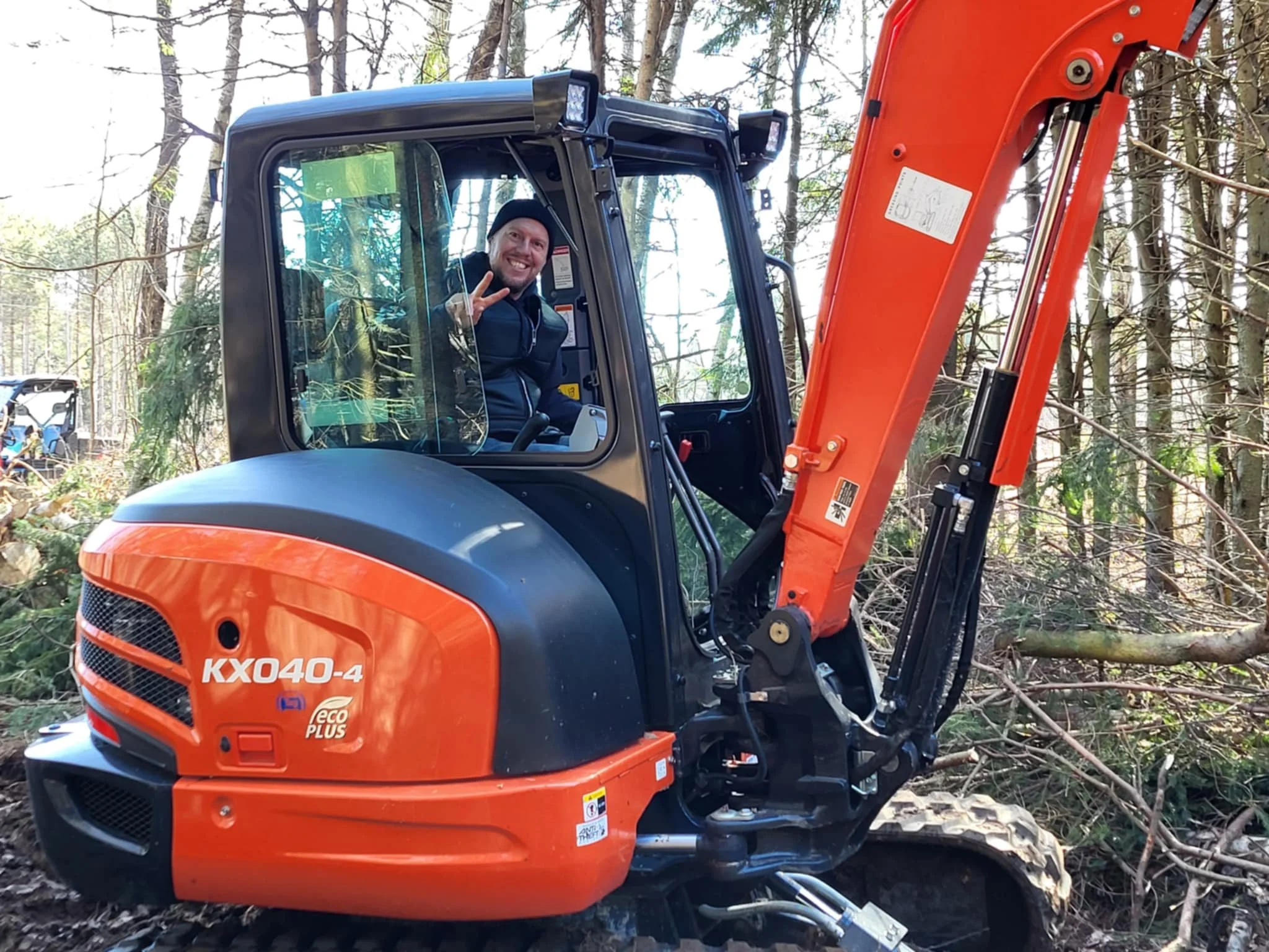 Une personne souriante, faisant un signe de paix, assise dans une machine excavatrice orange et noire dans une forêt.