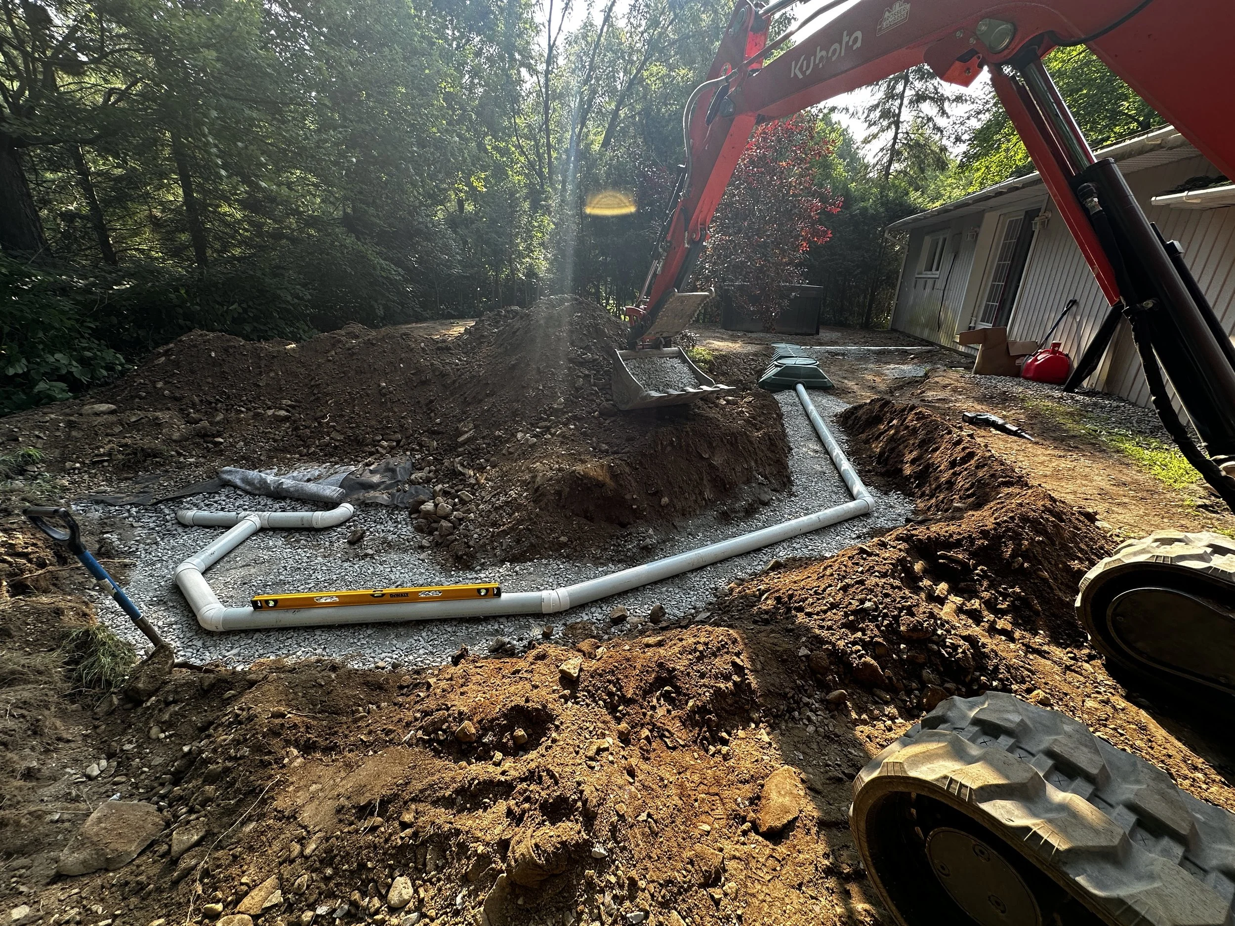 Travaux de plomberie en cours dans une cour arrière avec excavatrice, tuyaux en PVC, et un espace en construction entouré d'arbres, près d'une maison.