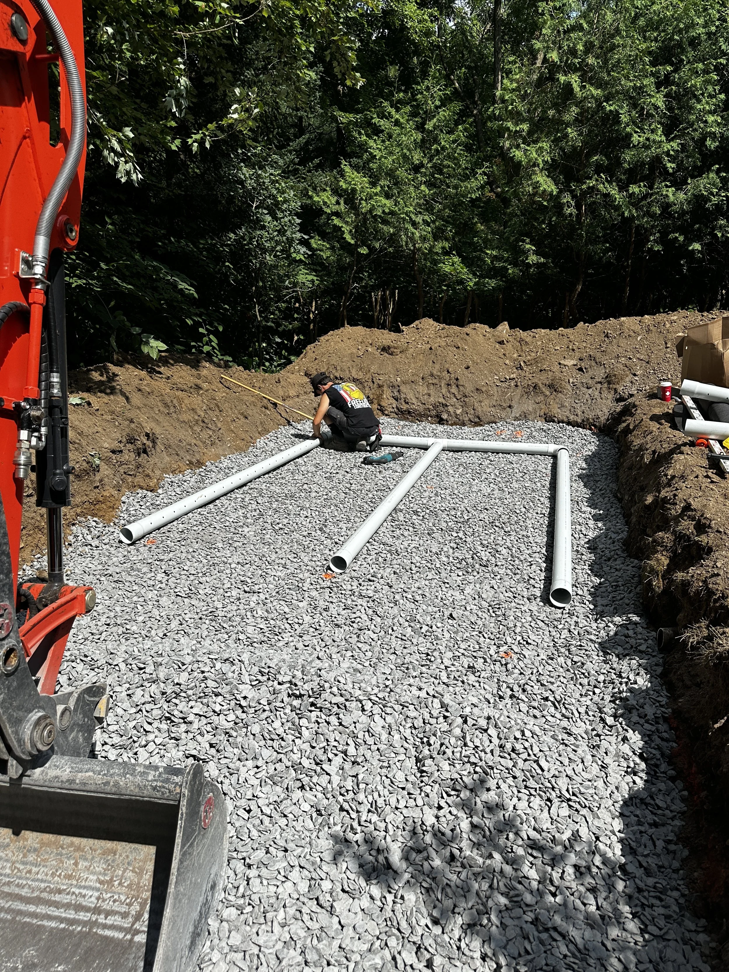 Travaux de pose de tuyaux dans un chantier en plein air, avec un ouvrier en train de les installer sur un sol en gravier, entouré d'une zone excavée et de verdure.