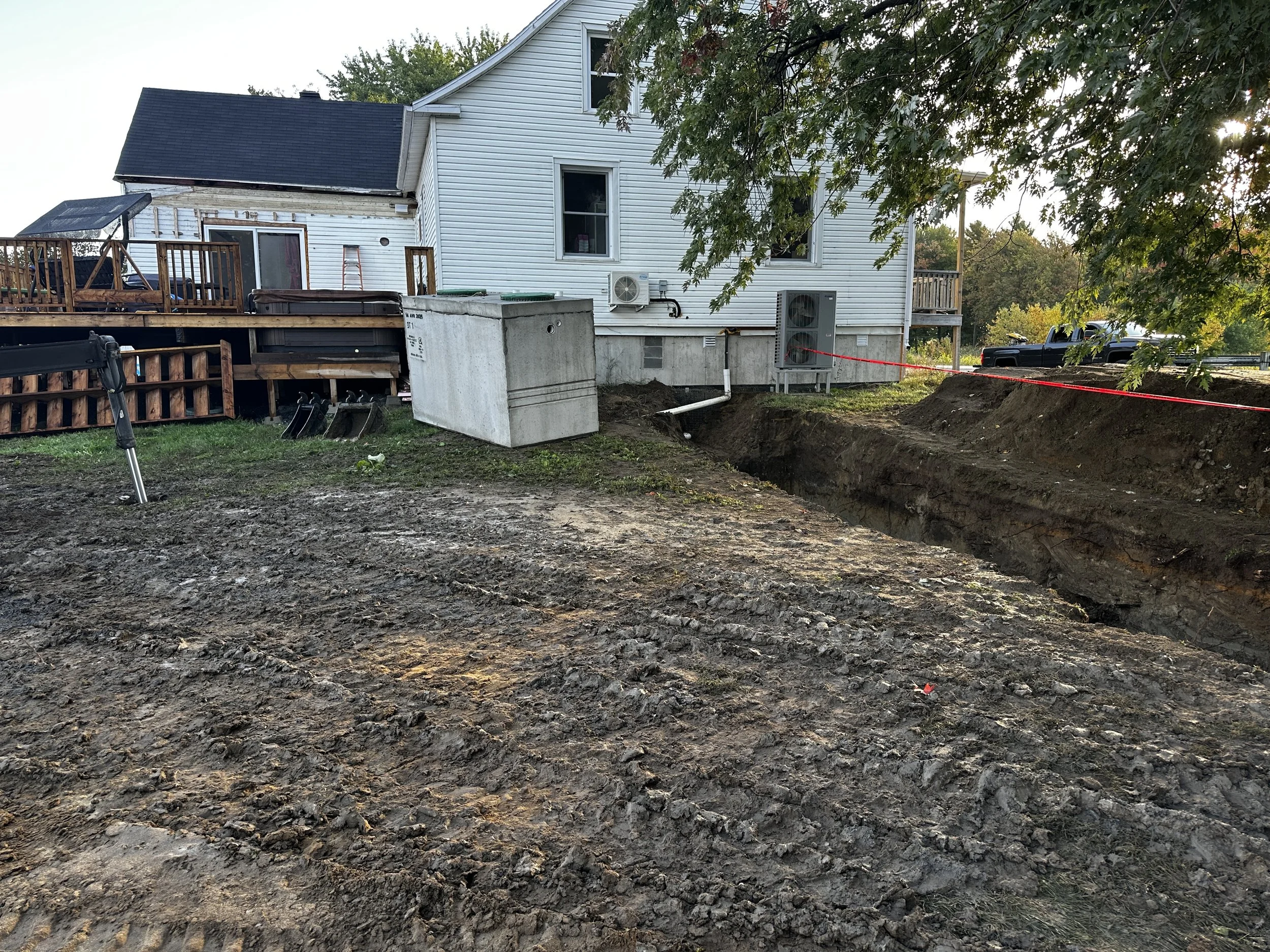 Une maison en cours de travaux avec une tranchée creusée dans le sol devant. La maison est blanche avec une terrasse en bois, et une petite plateforme en béton à l'avant. Des arbres verts encadrent l'image et le soleil brille à travers les branches.