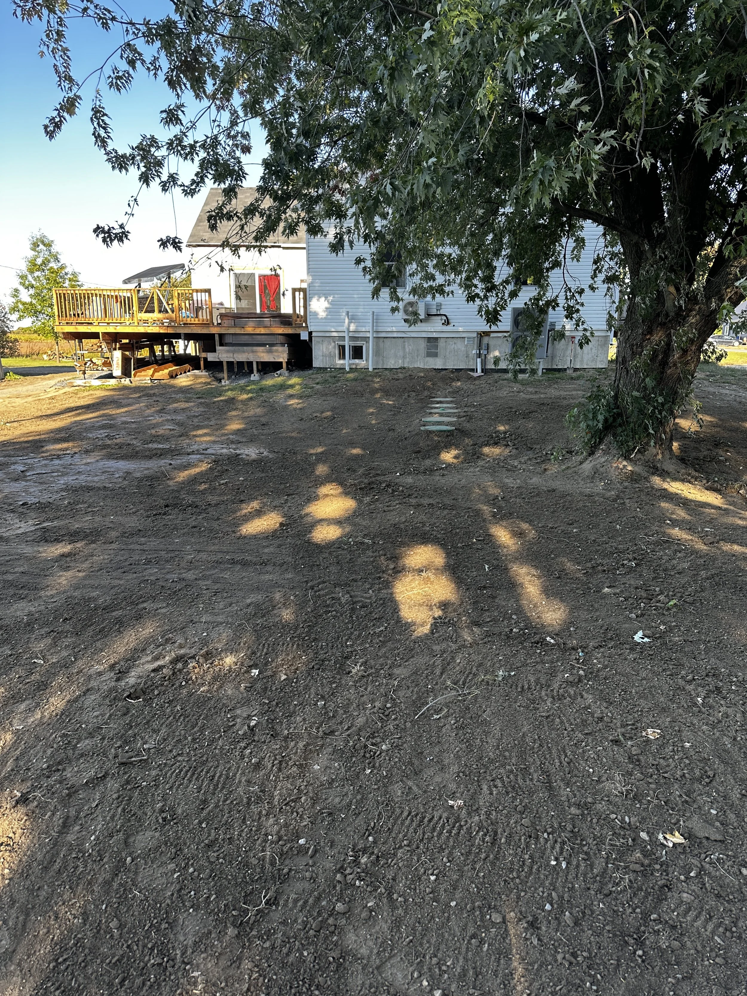 Maison en bois blanc sur une fondation en béton, située sur un terrain en terre dénudée, avec un arbre en premier plan et une véranda en bois avec une draperie rouge, dans un environnement extérieur en plein jour.