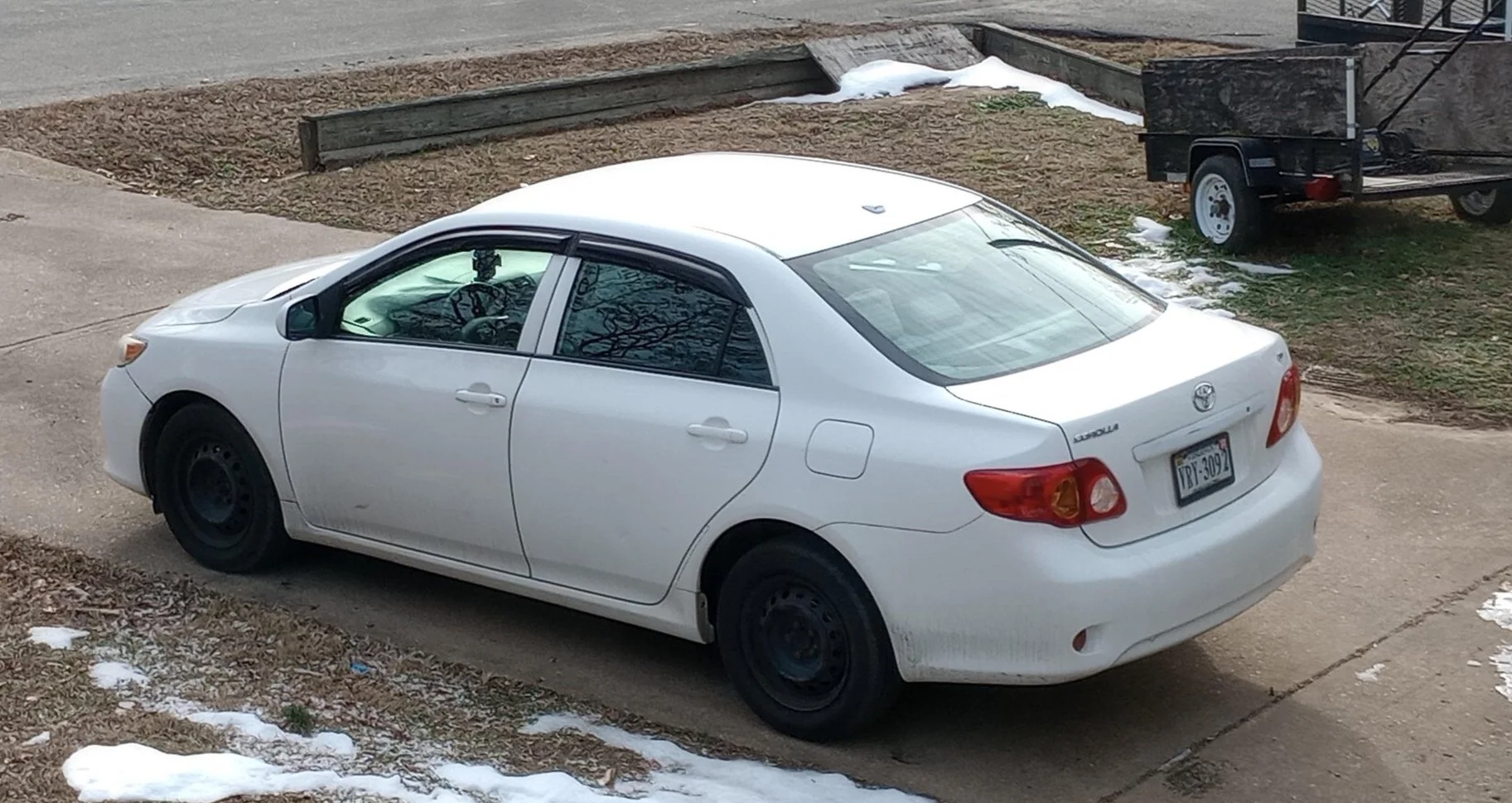 A white sedan parked on a concrete driveway with patches of snow on the ground, a trailer parked on grass nearby, and a wooden planter box behind the car.